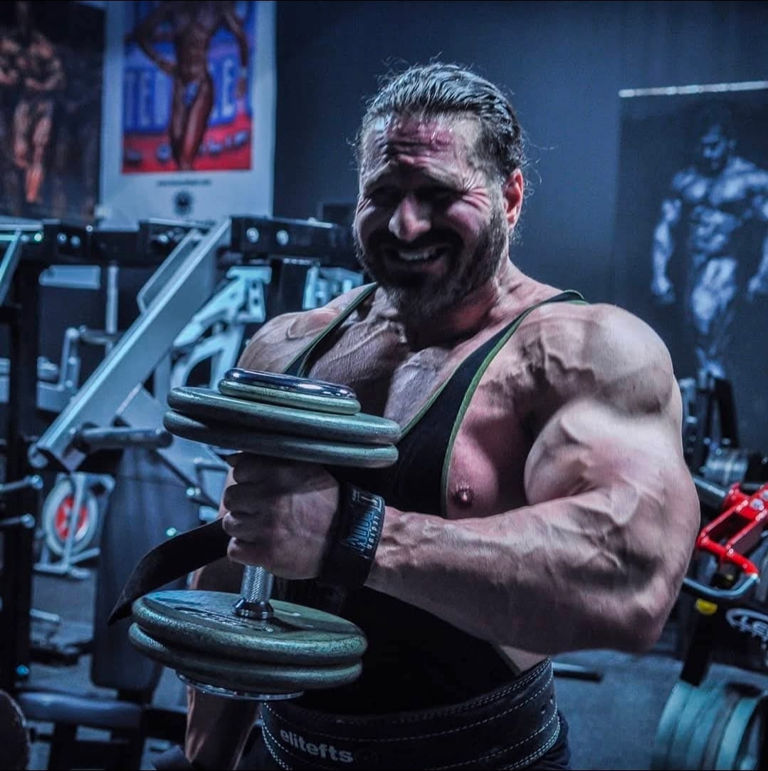 Muscular man lifting dumbbells in a gym, smiling, with gym equipment and posters in the background.
