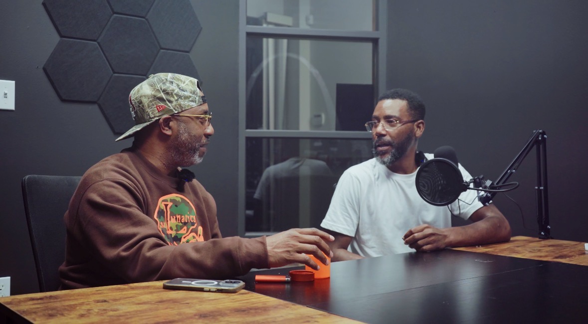 Two men sit at a table in a recording studio, engaging in conversation with a microphone nearby.