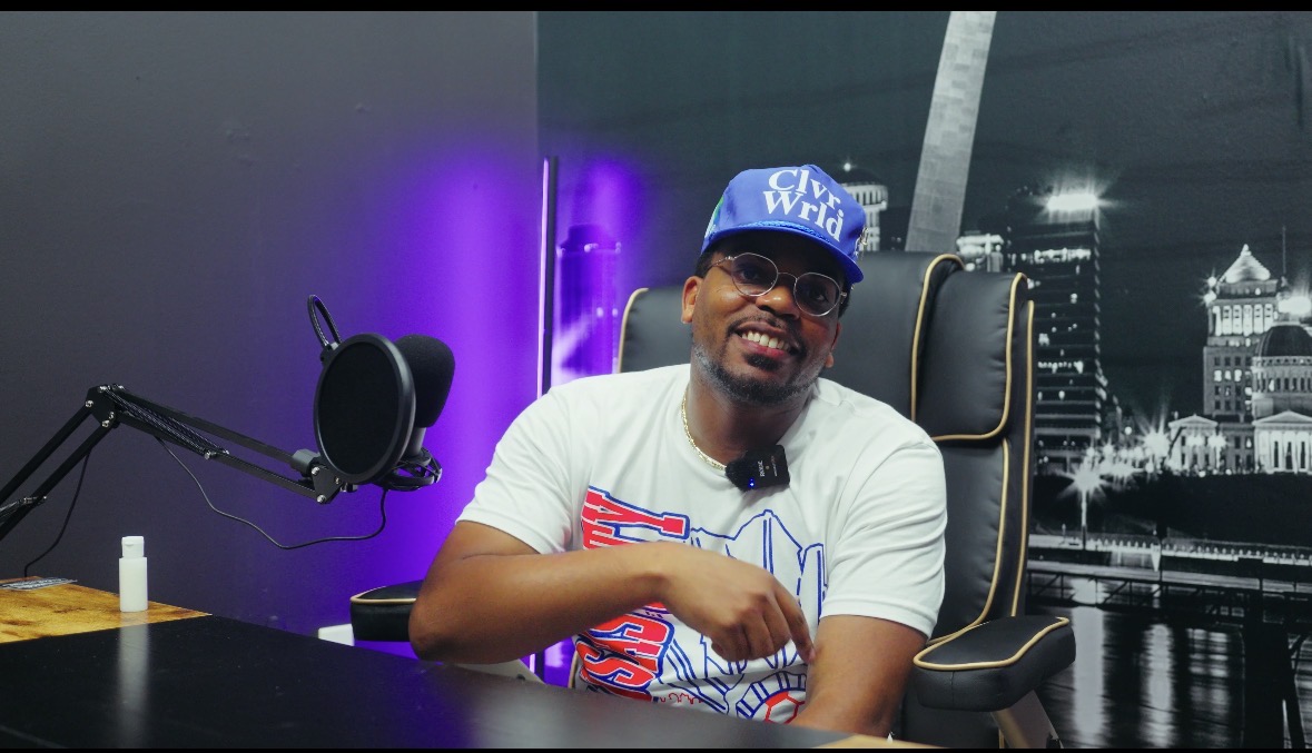 Man wearing a blue cap and sunglasses sitting in a chair in a studio with a purple and black background.