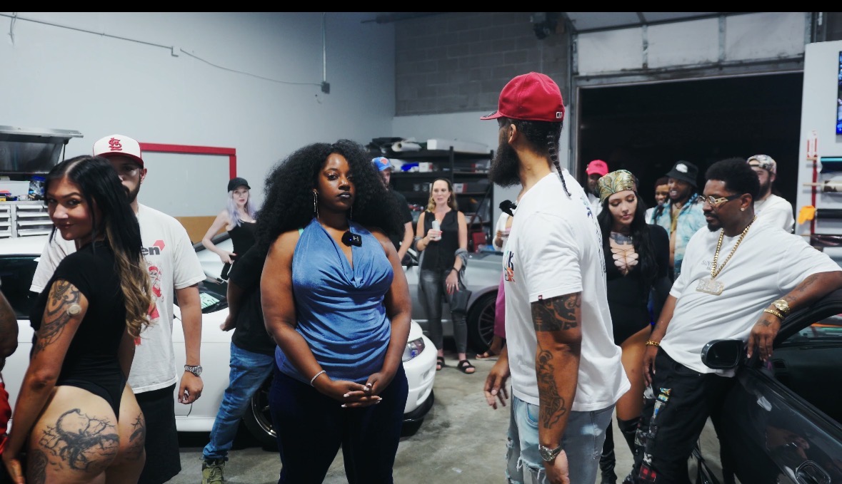 Group of people in an indoor space, some talking and others standing near cars, with a woman in a blue top in the center.