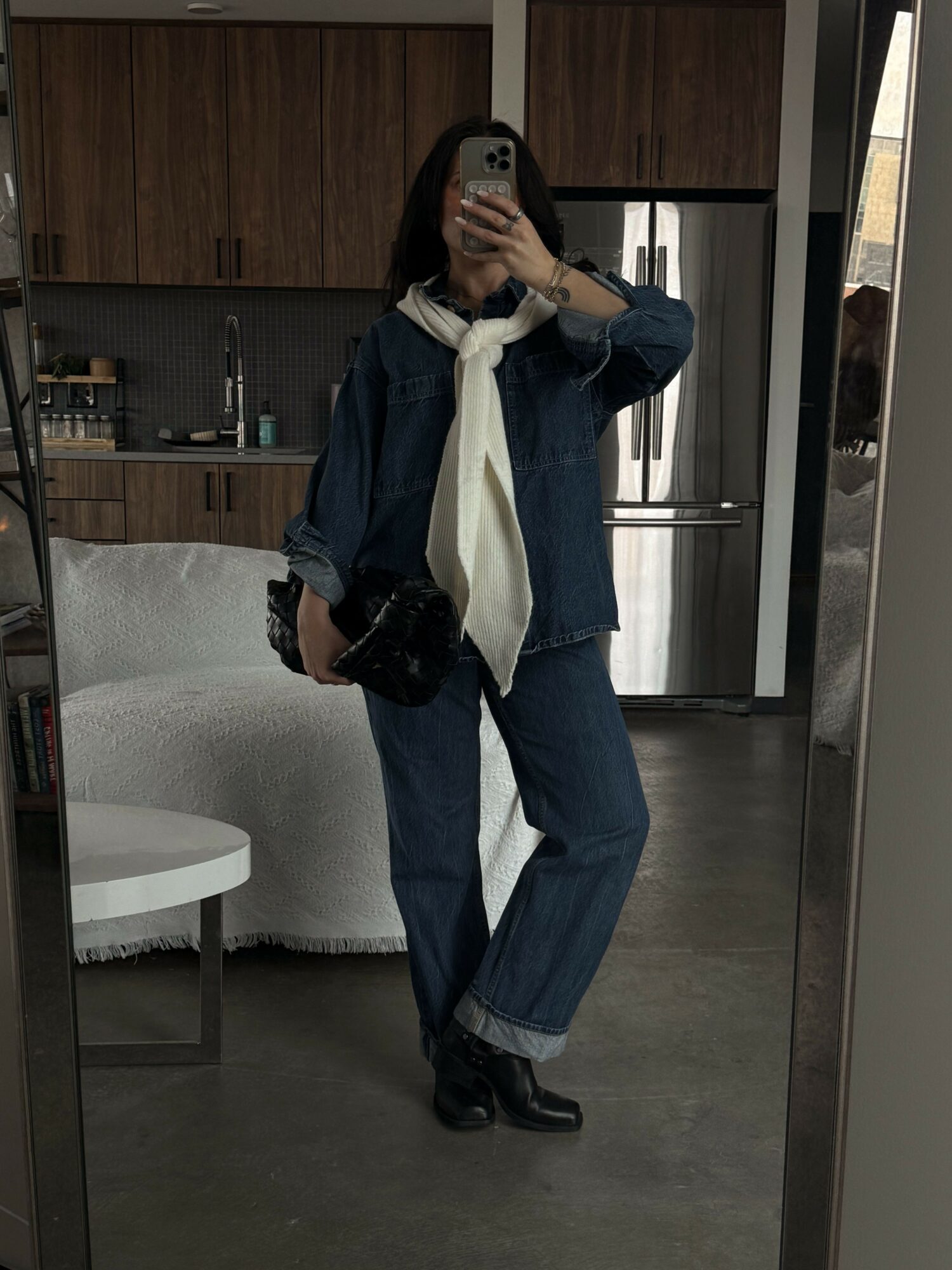 Person taking a mirror selfie in a room with wooden cabinets, wearing a dark jacket, white shirt, and jeans, holding a phone.