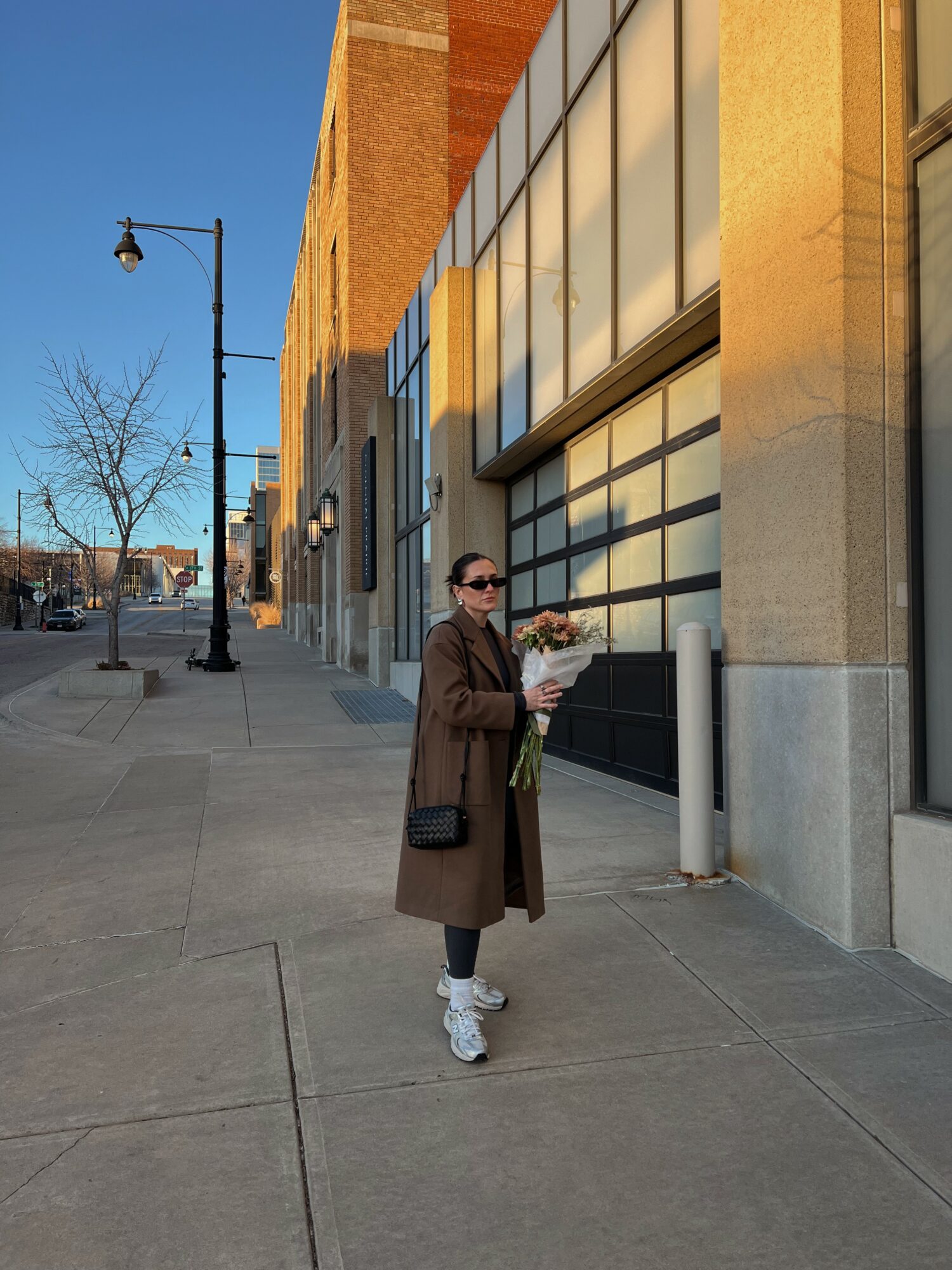 Person in brown coat holding flowers on sidewalk near building with glass windows, streetlamp, and trees in background.