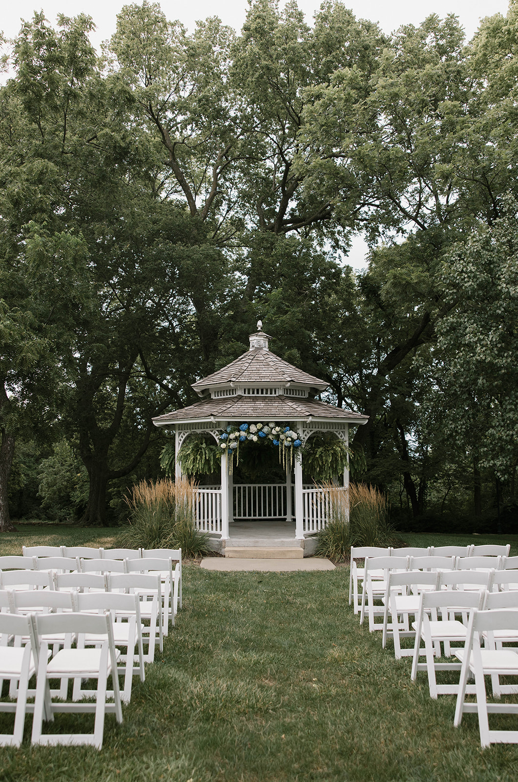 Gazebo with white railings and a shingled roof, surrounded by trees and white chairs on grass, outdoor setting.