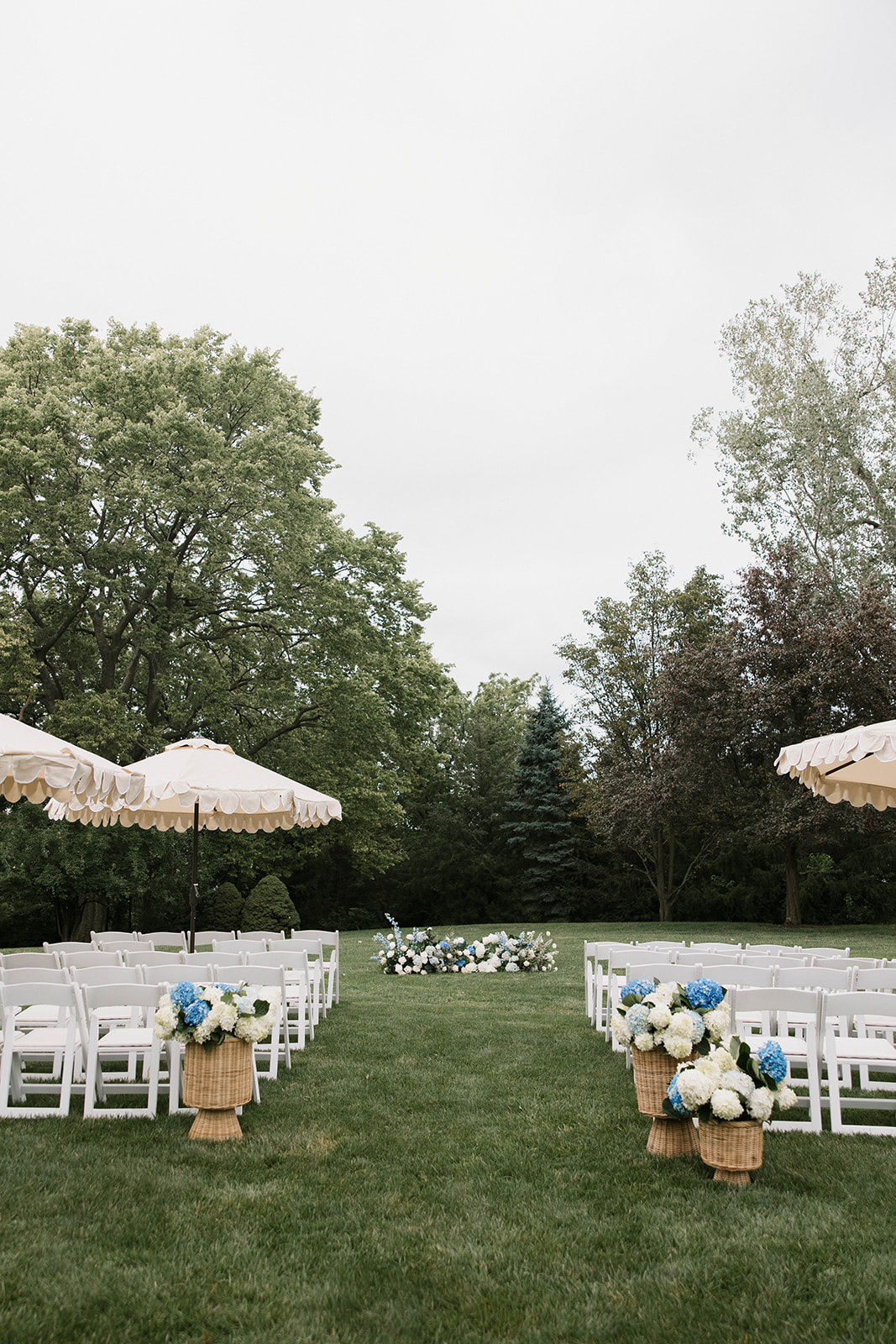 Outdoor wedding setup with white chairs, floral arrangements, and umbrellas on a grassy area, trees in the background.
