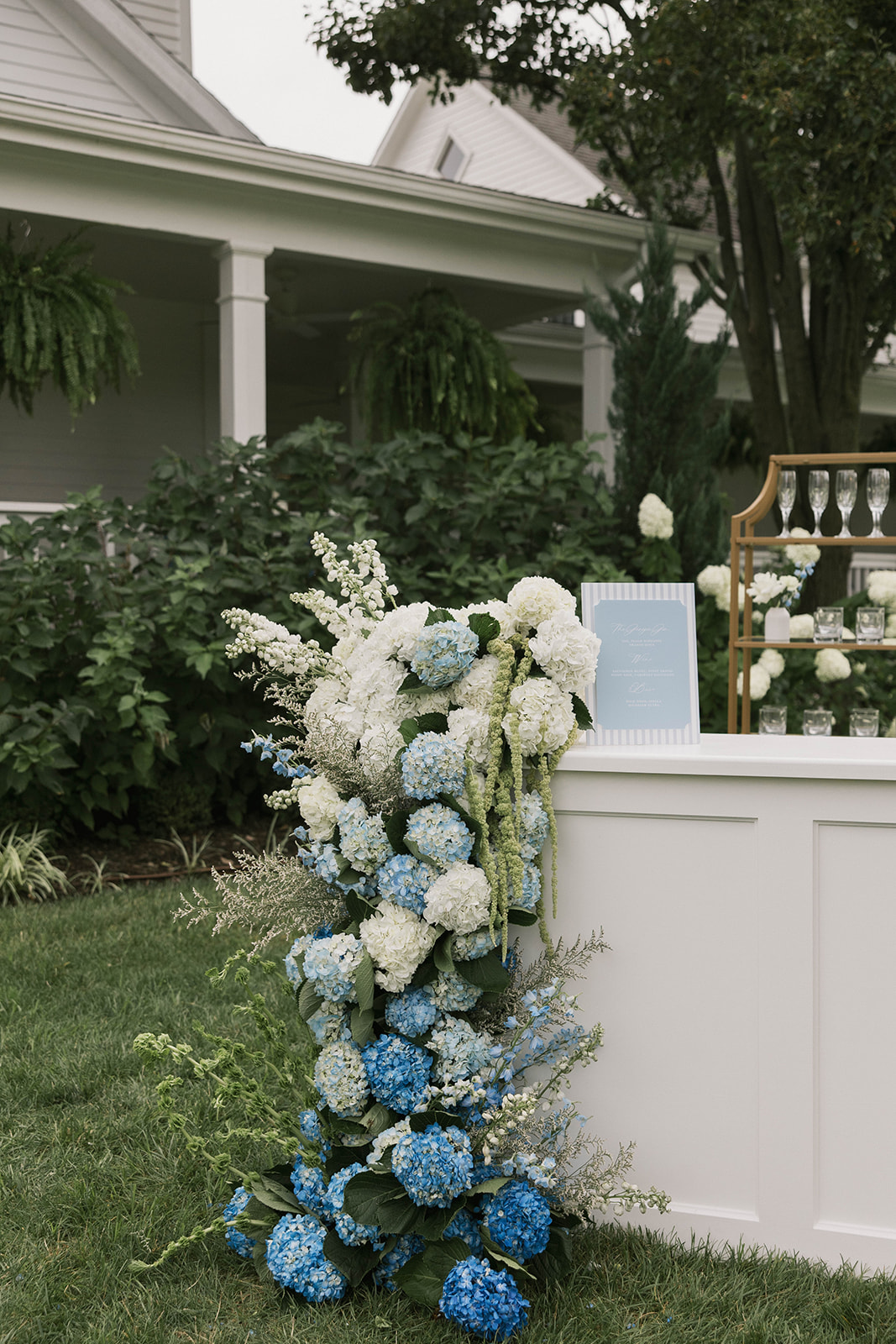 Floral arrangement with white and blue flowers on grass in front of a white table, house, and trees in background.
