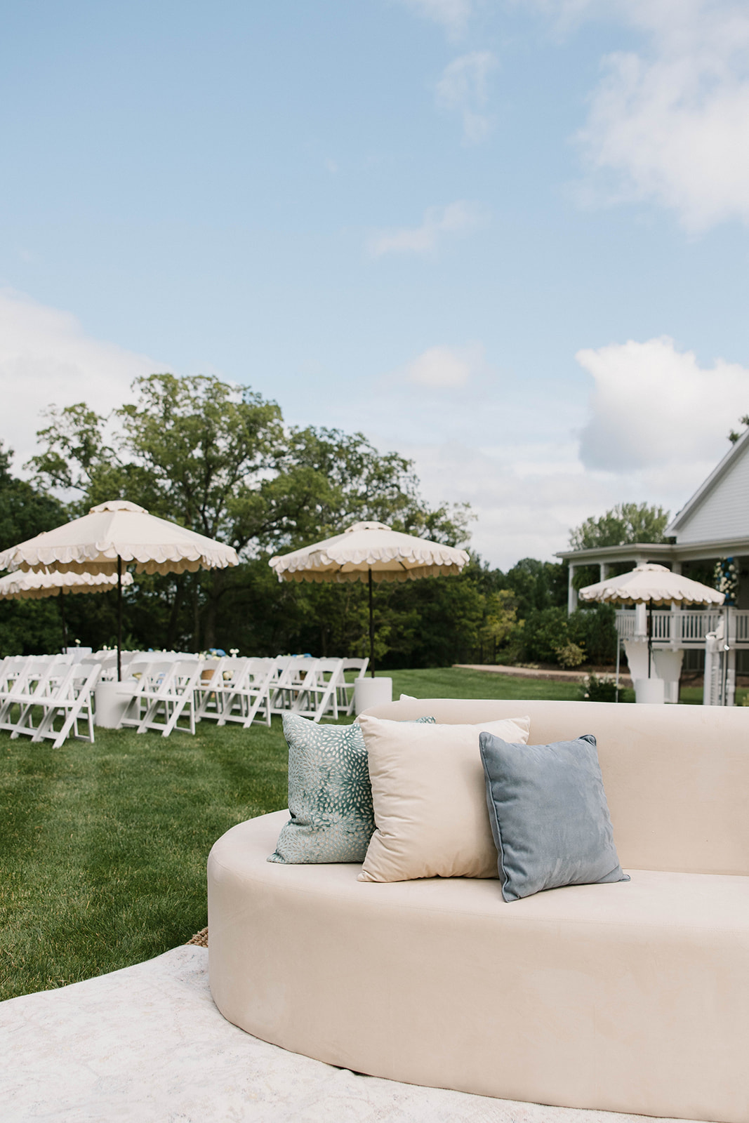 Outdoor seating area with white chairs, umbrellas, and a curved beige sofa with pillows on a grassy lawn under a blue sky.