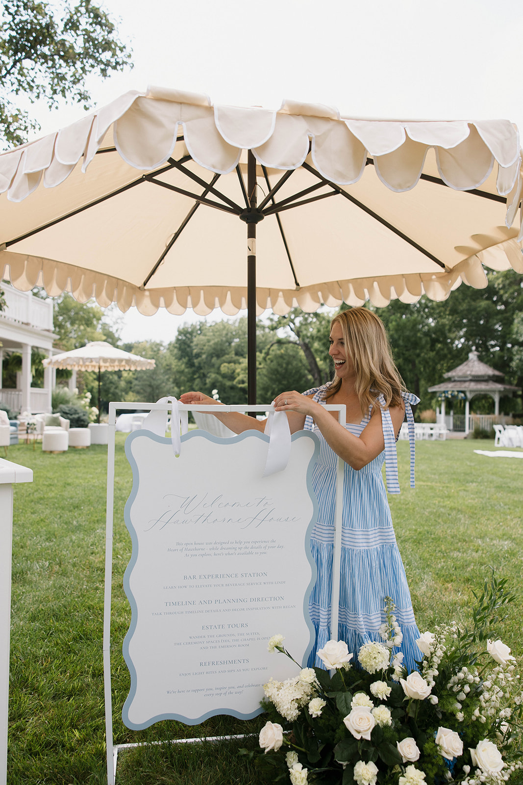 Young girl in a blue dress standing outdoors near a large white sign with flowers and a patio umbrella in the background.