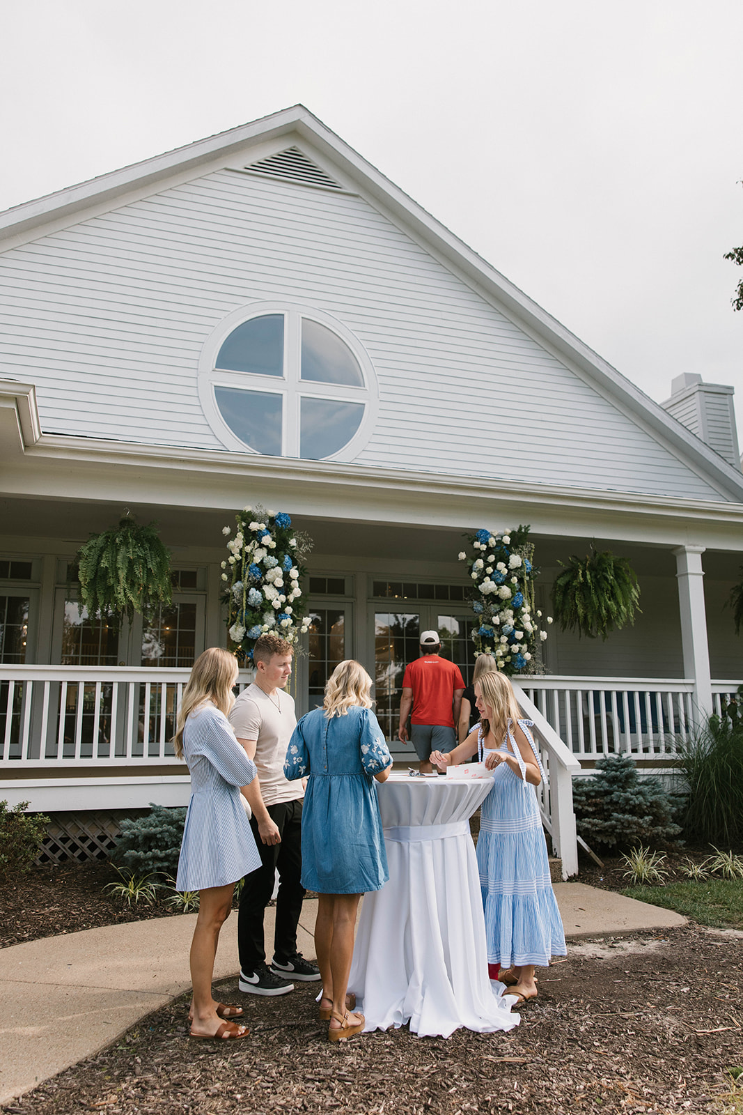 Group of people gathered outside a white house with a circular window, decorated with flowers, during daytime.
