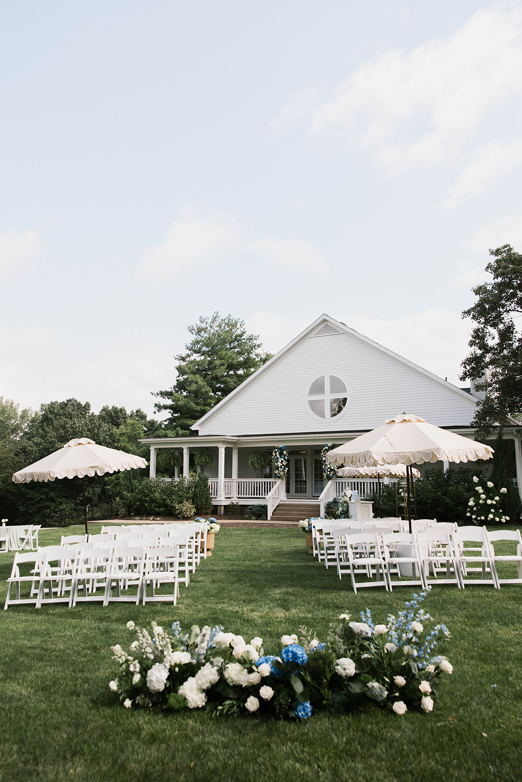 Outdoor wedding setup with white chairs, umbrellas, and floral arrangements in front of a white building with a circular window.
