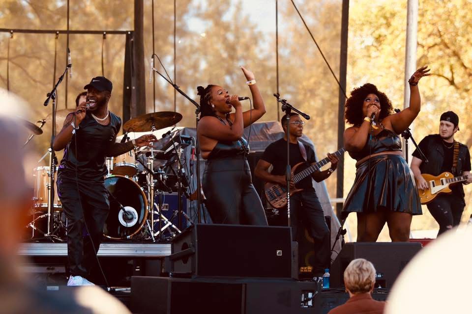 Group of five musicians performing on stage outdoors with trees in the background, singing and playing instruments.