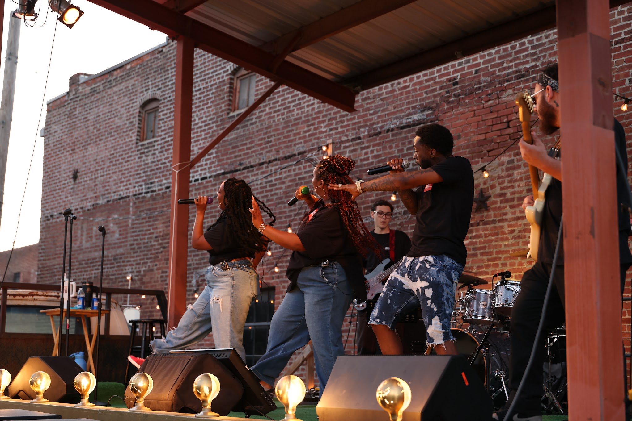 Group of people dancing and playing instruments on stage outdoors, brick wall background, stage lighting, and musical equipment visible.