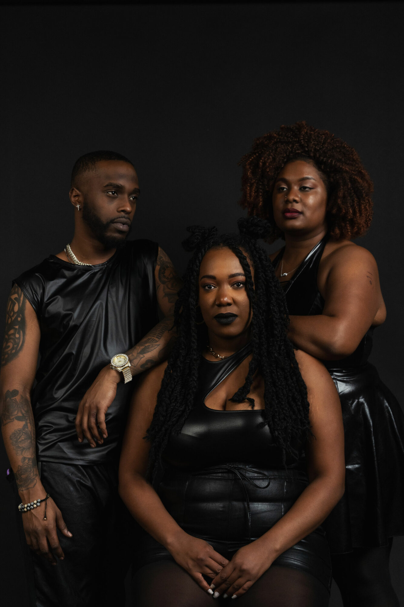 Three women pose together against a dark background, wearing black outfits, with serious expressions.