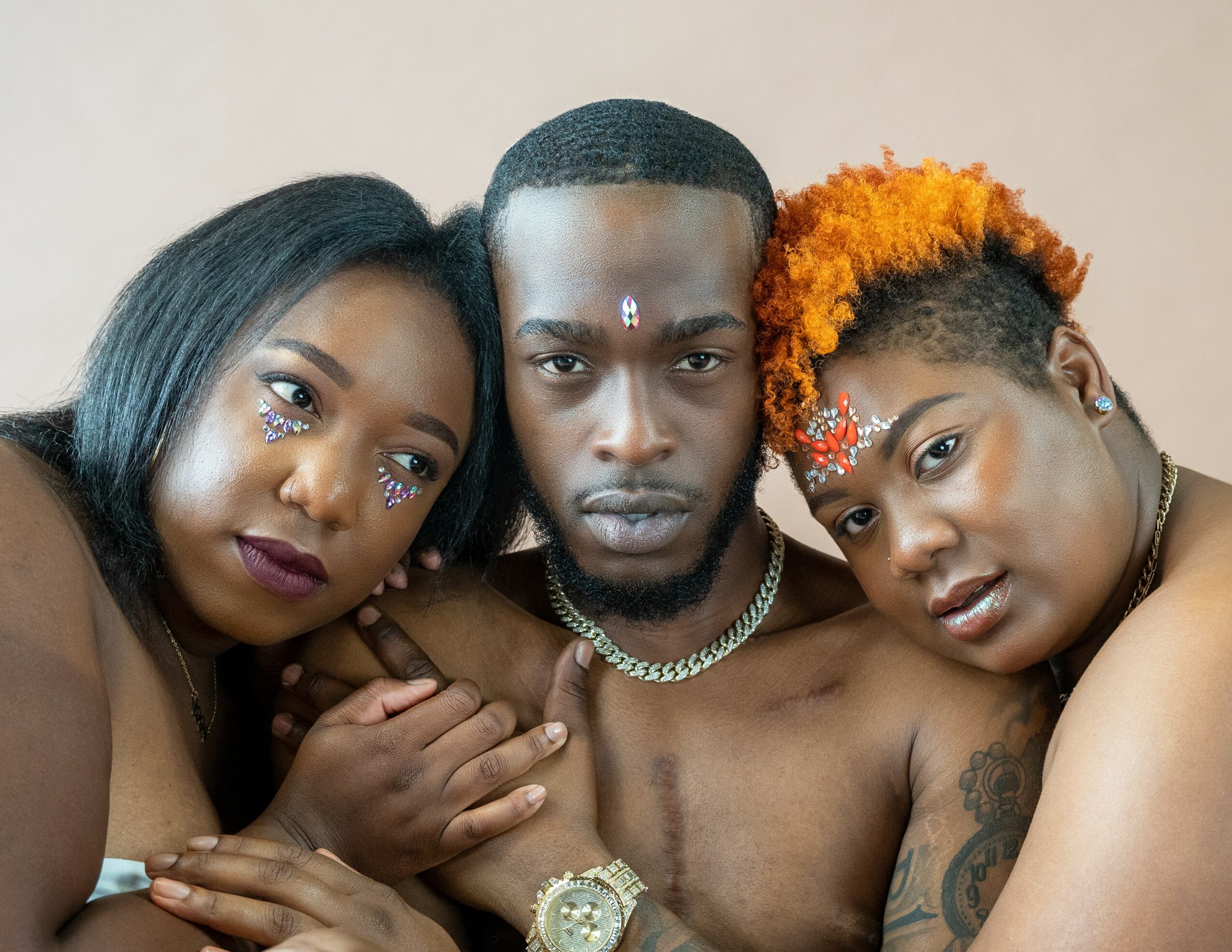Three people with face paint and jewelry close together, looking at the camera, against a plain background.