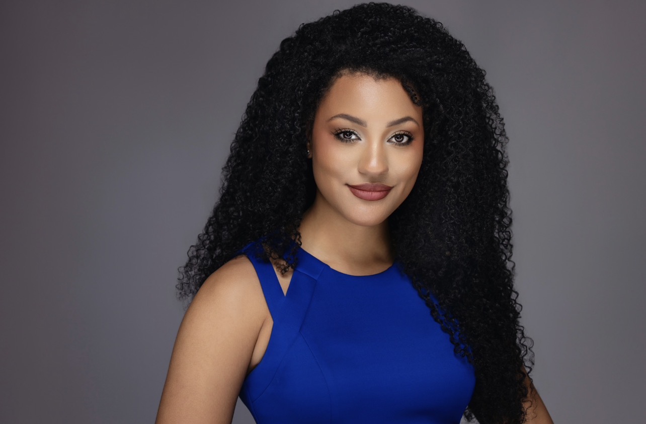 Portrait of a woman with long curly black hair wearing a blue sleeveless top, smiling against a gray background.