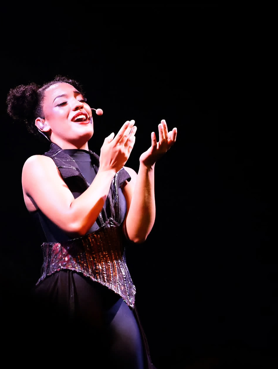 A woman on stage smiling, clapping her hands, wearing a black outfit with a glittery belt, against a black background.
