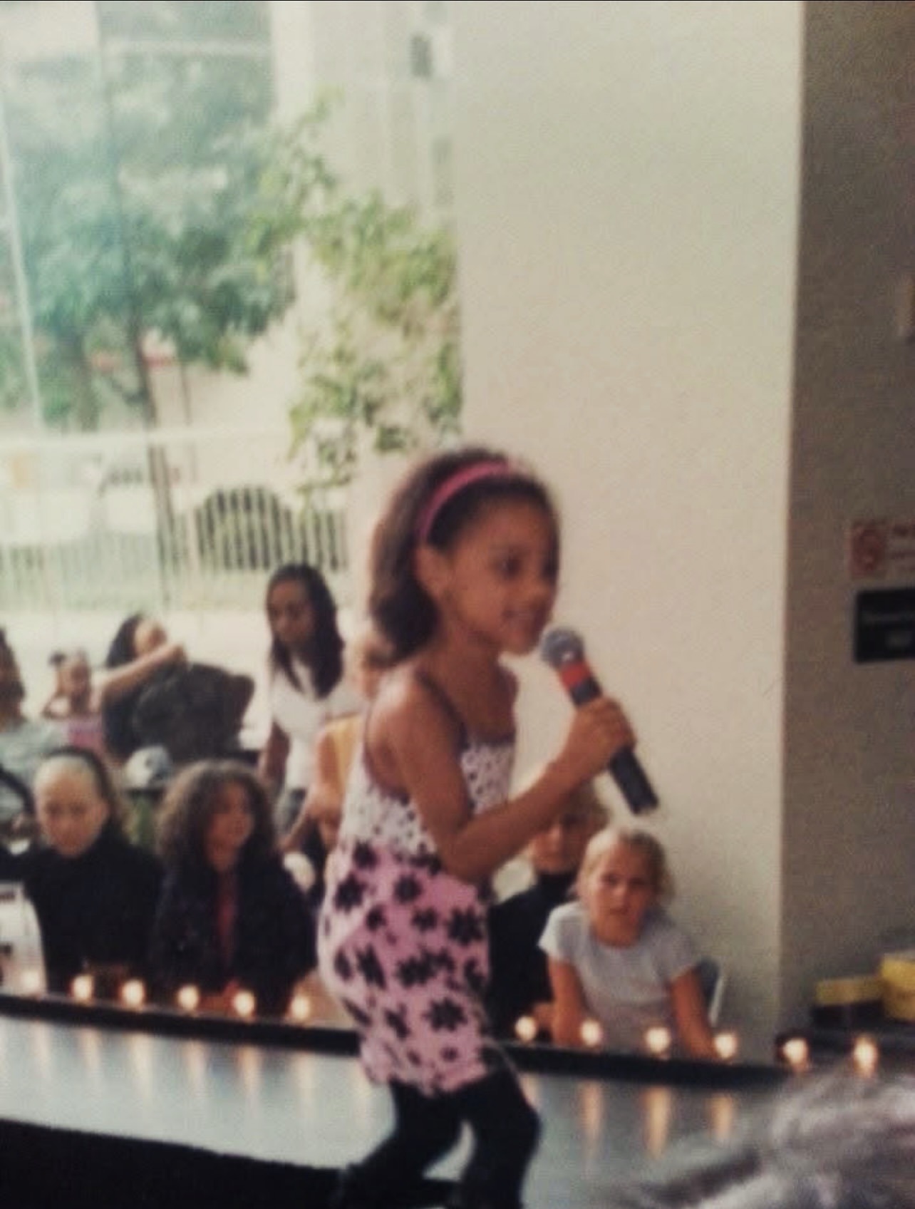 Young girl holding a microphone, standing in front of seated children indoors, with large windows showing trees outside.