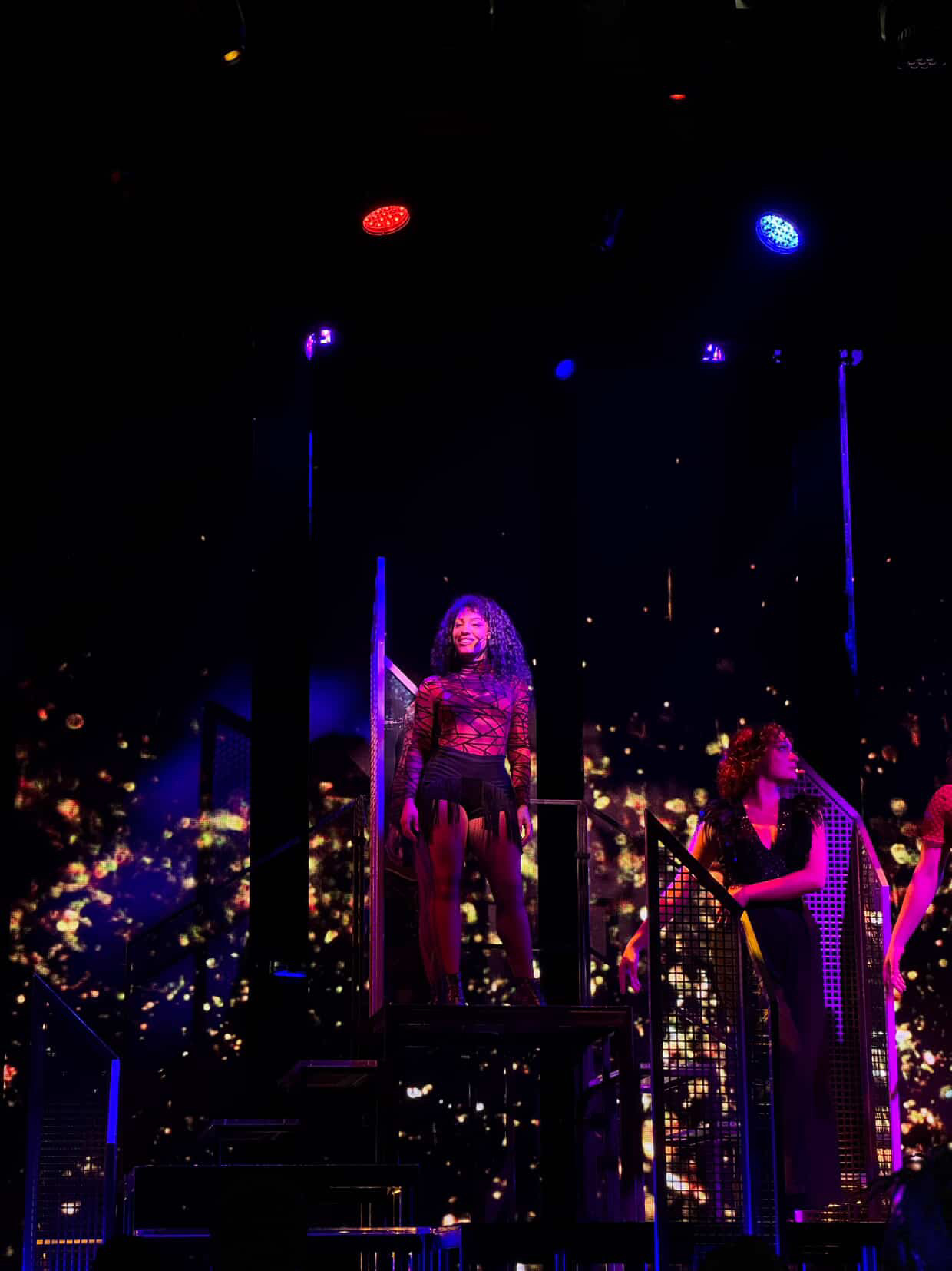 Performer on stage with purple lighting, standing on a platform, with another person nearby, cityscape background with lights, night scene.
