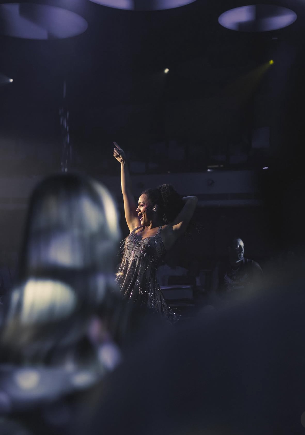 A performer on stage with one arm raised, wearing a sparkly outfit, in a dark setting with stage lights.