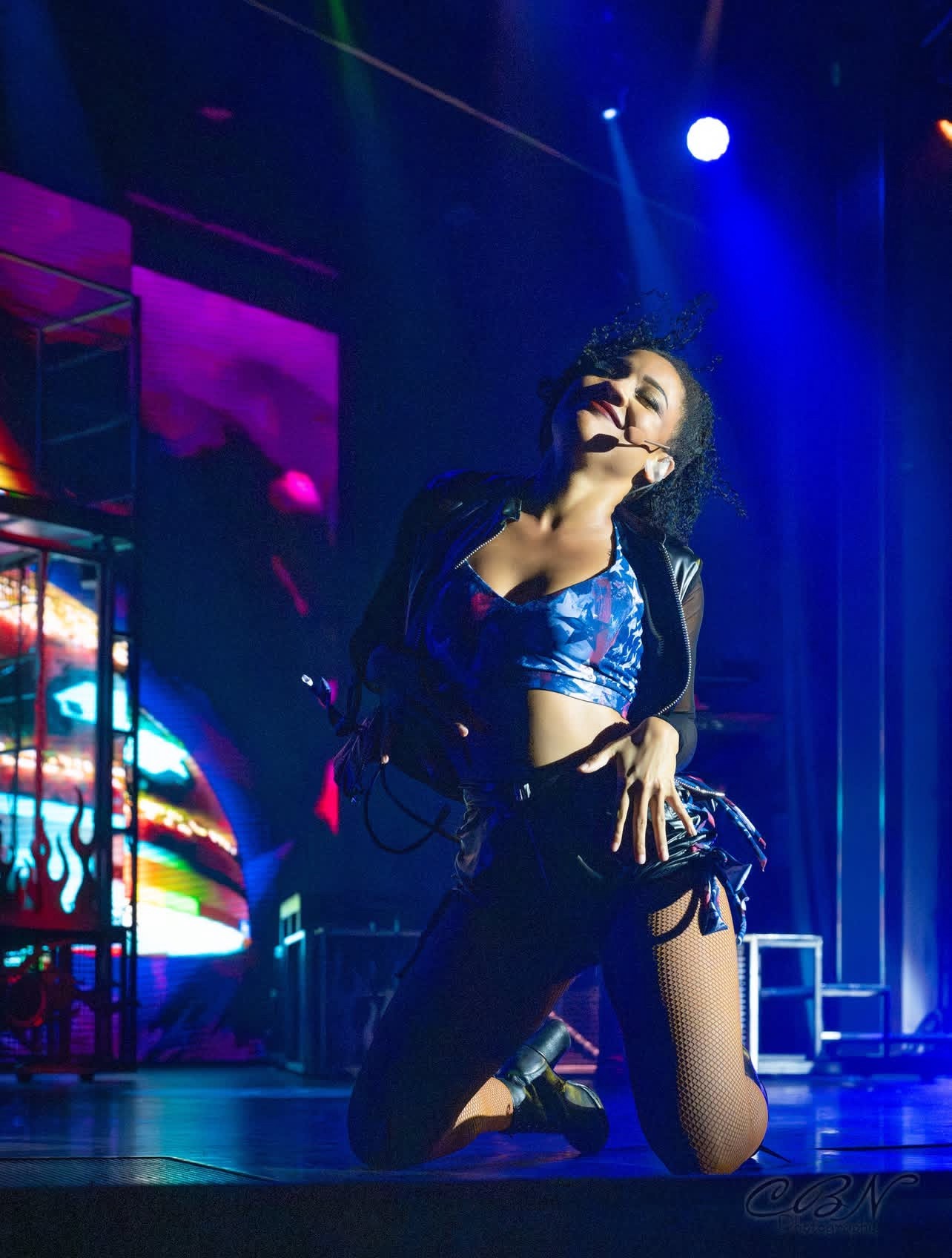 Kneeling performer on stage with colorful background lighting, wearing a shiny blue outfit, in a dynamic pose.
