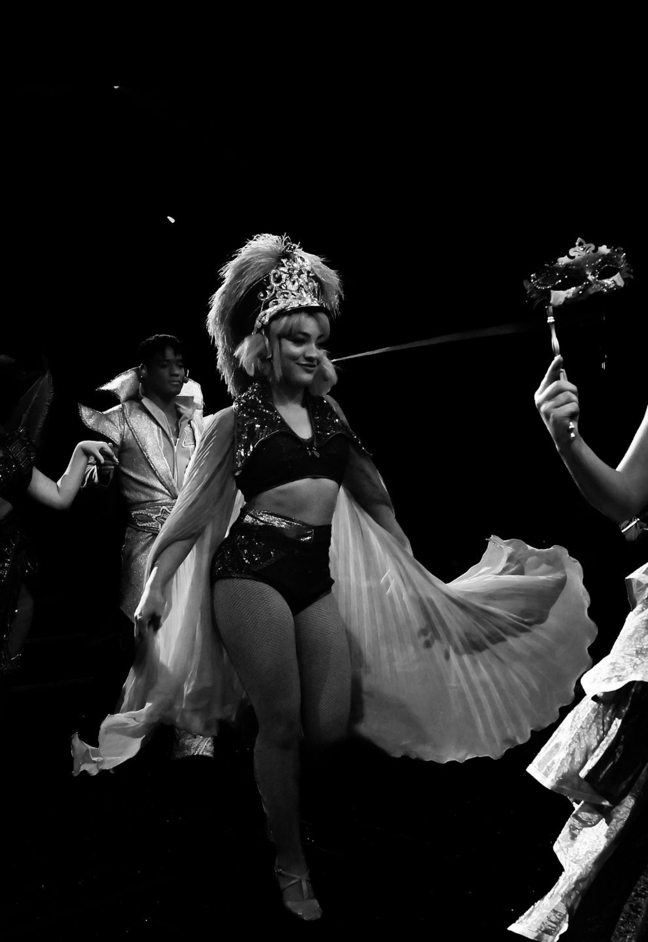 Performer in costume with a feathered headdress and flowing cape on stage, holding a bouquet, with other performers nearby.