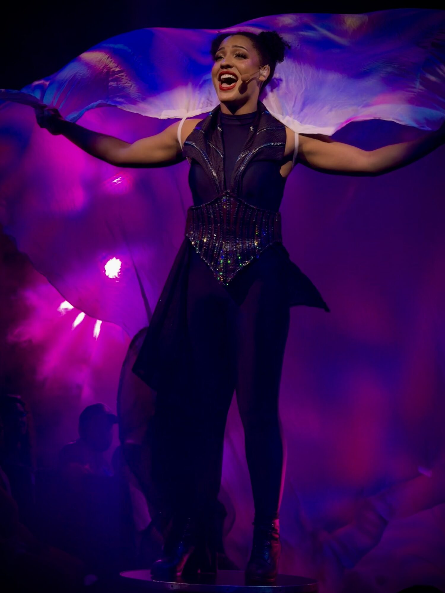 Performer with arms outstretched, smiling on stage with colorful background lighting.