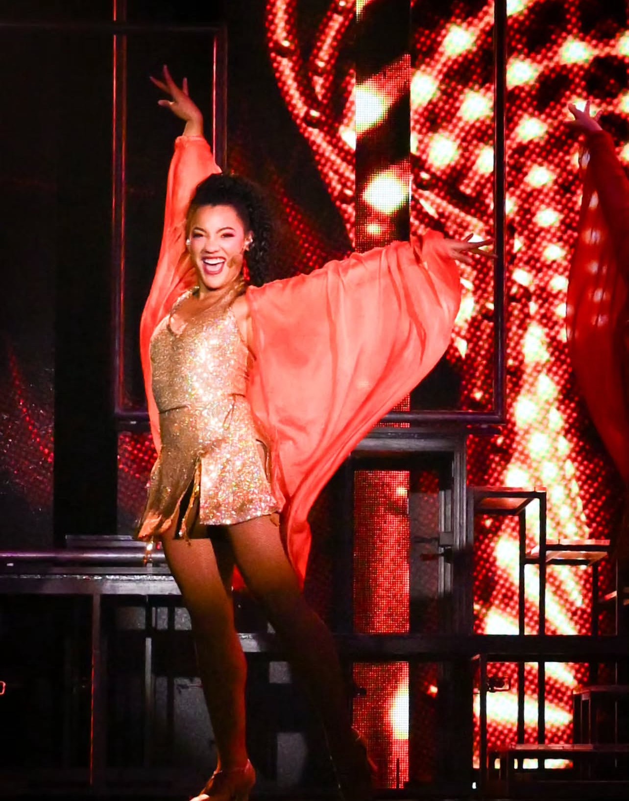 A woman in a gold dress and flowing red cape dances on stage with a bright, colorful background.