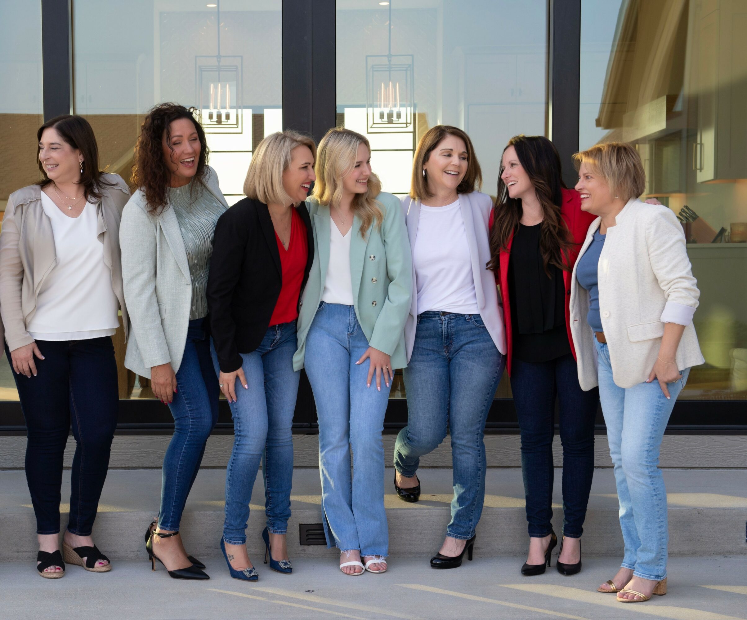 Seven women standing together outside a building, smiling and talking, with large windows behind them.