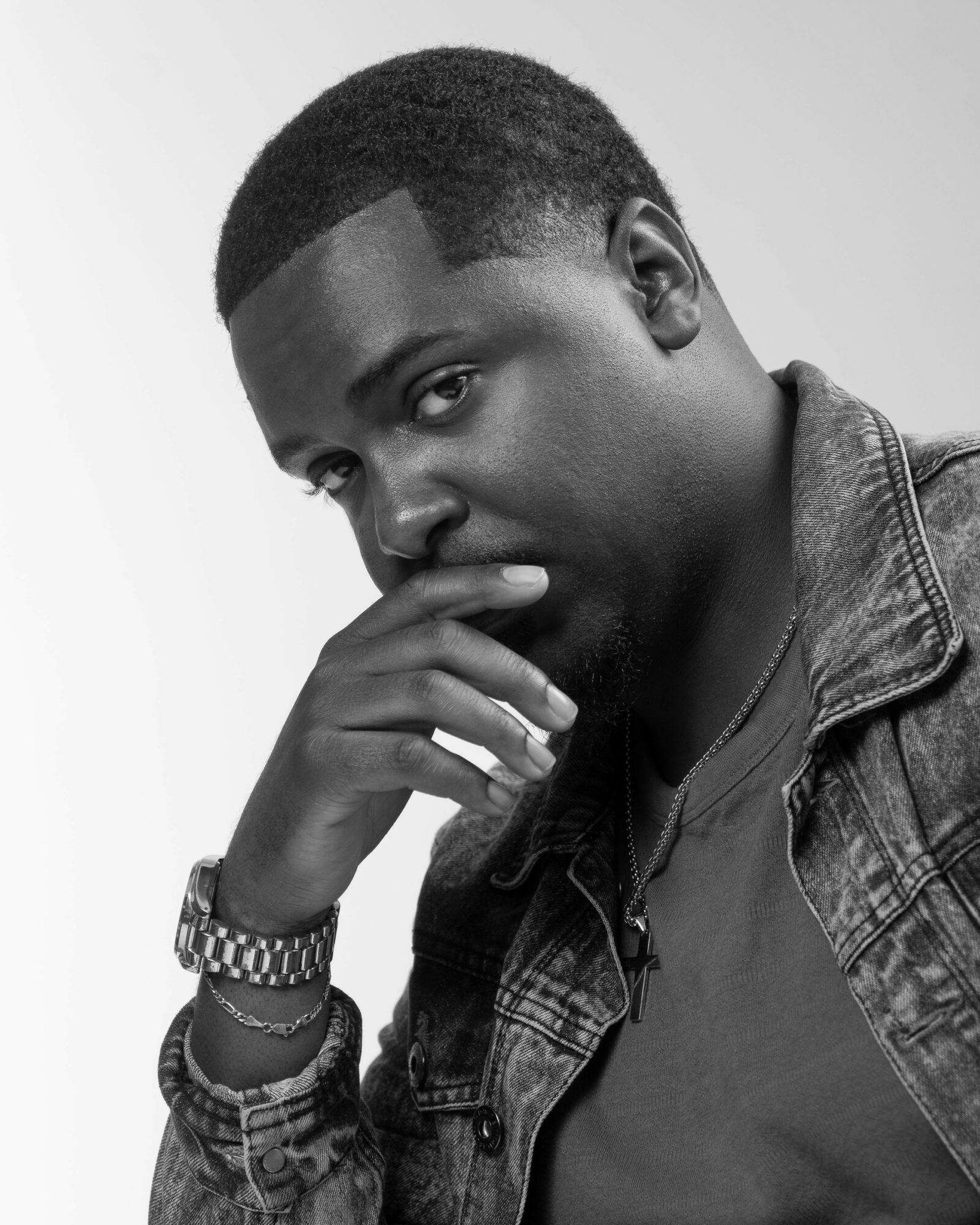 Portrait of a man with short hair, wearing a denim jacket and watch, resting his chin on his hand, looking thoughtful.