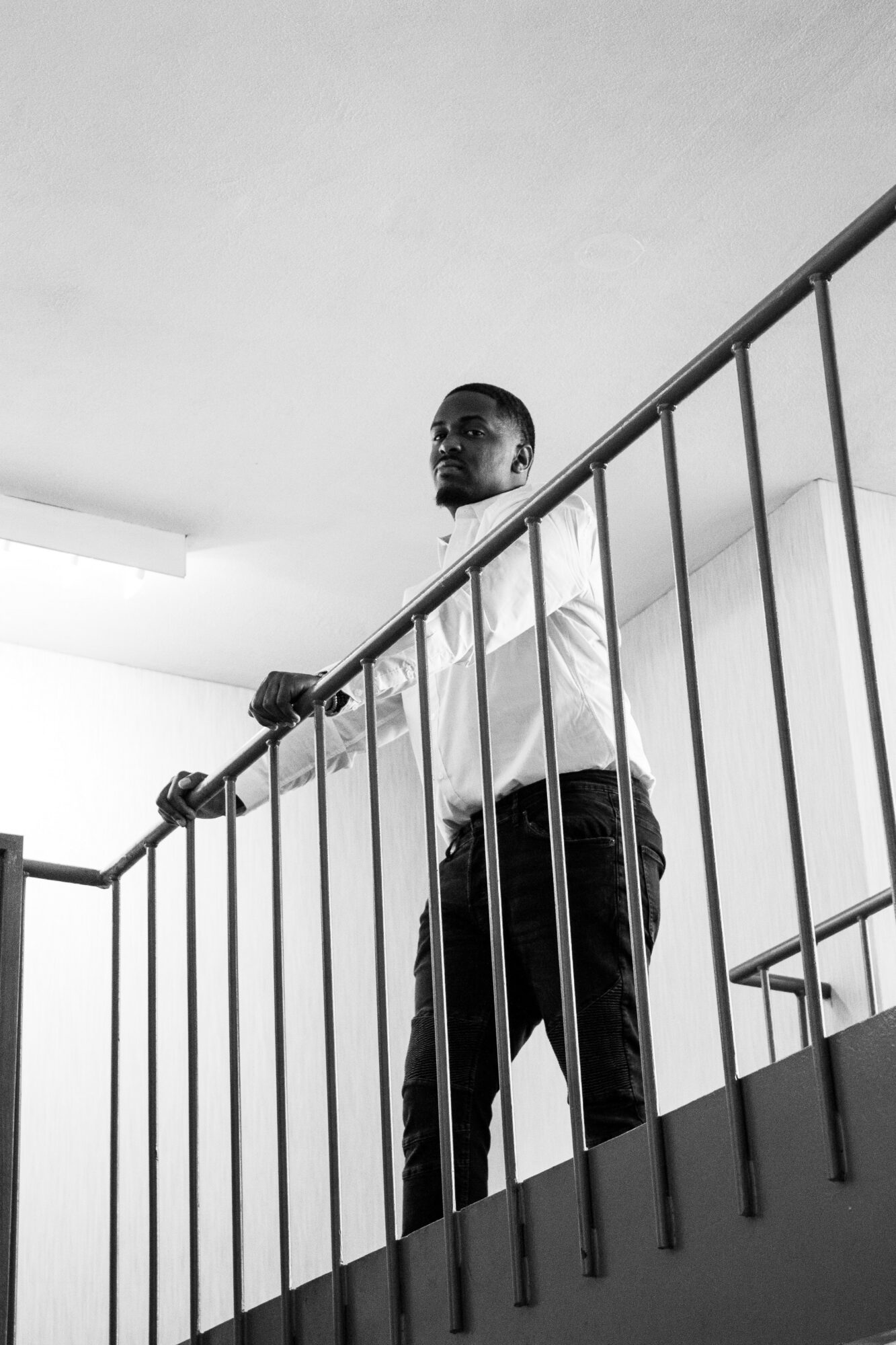 Person standing behind railing, looking over, in a room with plain ceiling and wall, black and white photo.