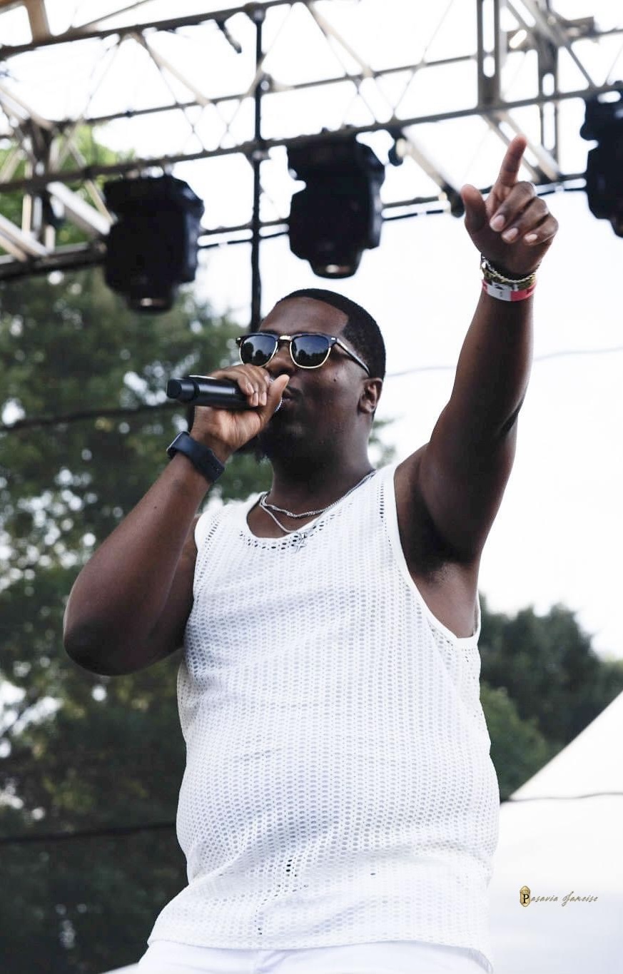 Male performer wearing sunglasses and a white sleeveless shirt, holding a microphone, raising one hand, outdoors stage setting