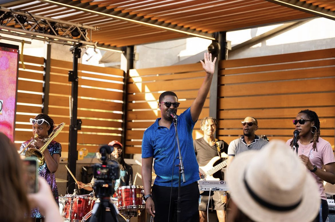 Man in blue shirt raising hand on stage with musicians and audience, wooden backdrop, outdoor setting.