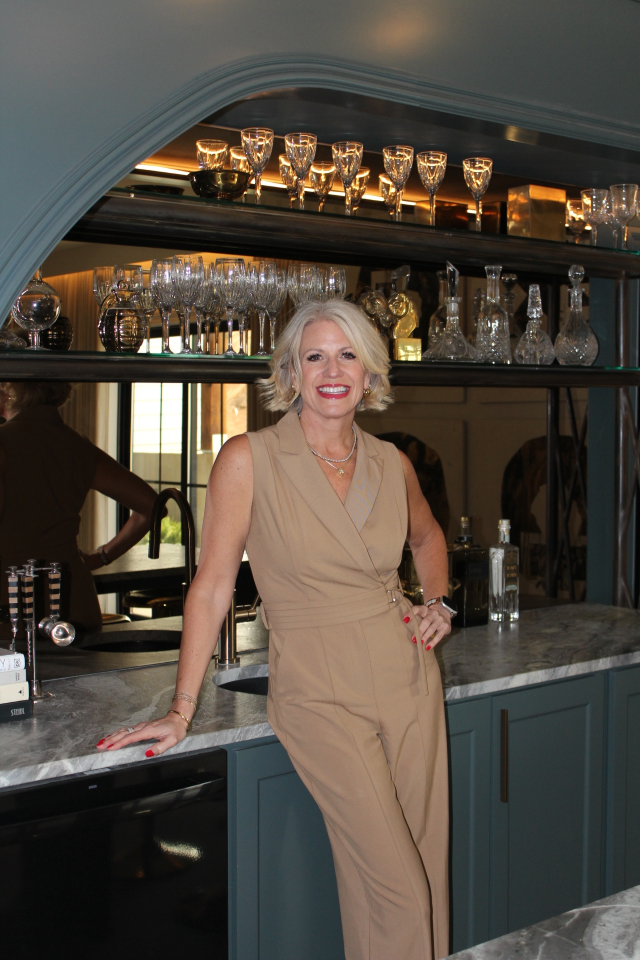 Woman with blonde hair in beige outfit standing at bar counter, smiling, glasses and bottles behind her.