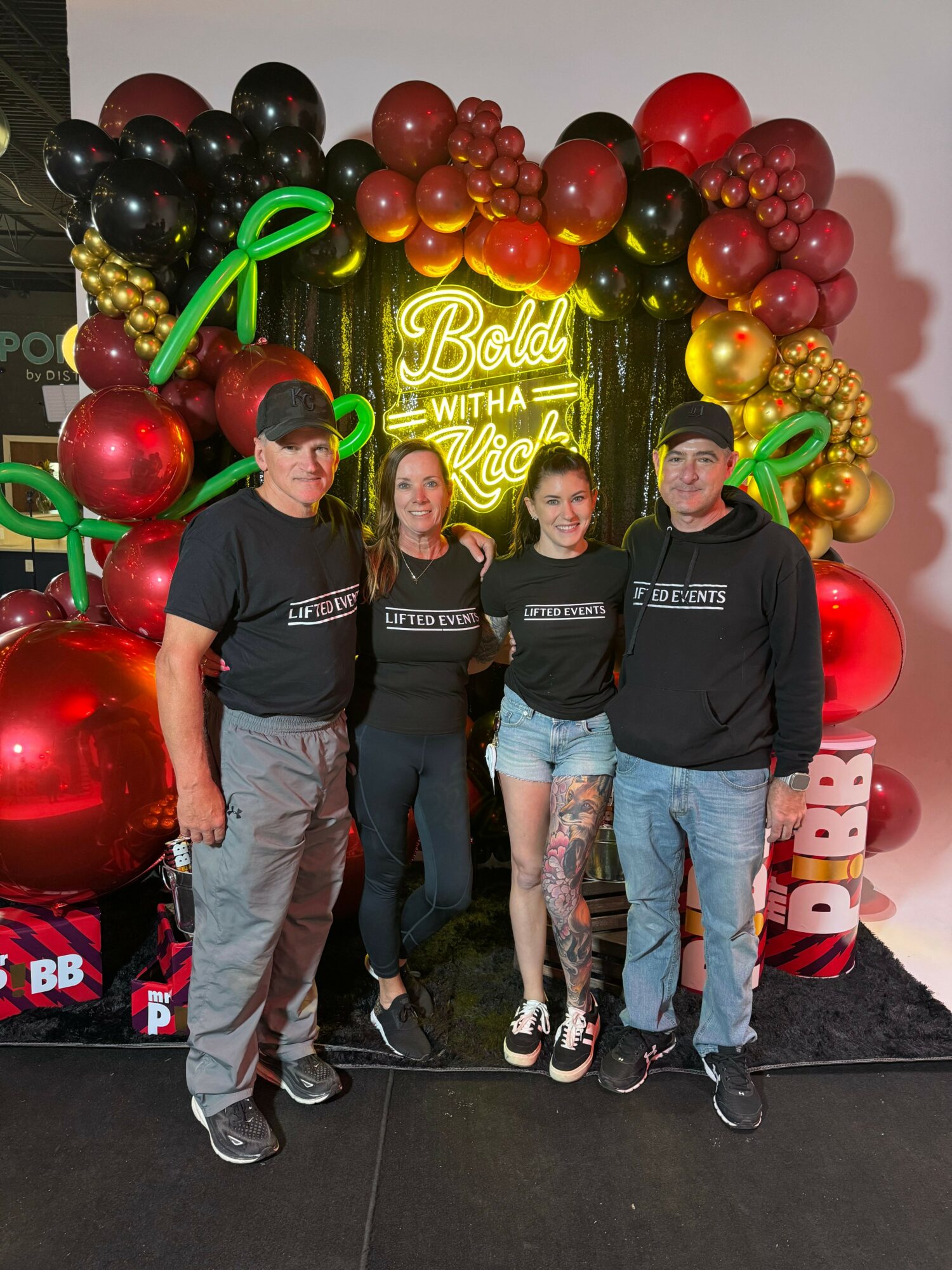 Three people standing in front of a balloon display with a neon sign, balloons in red, gold, and black, and a black background.