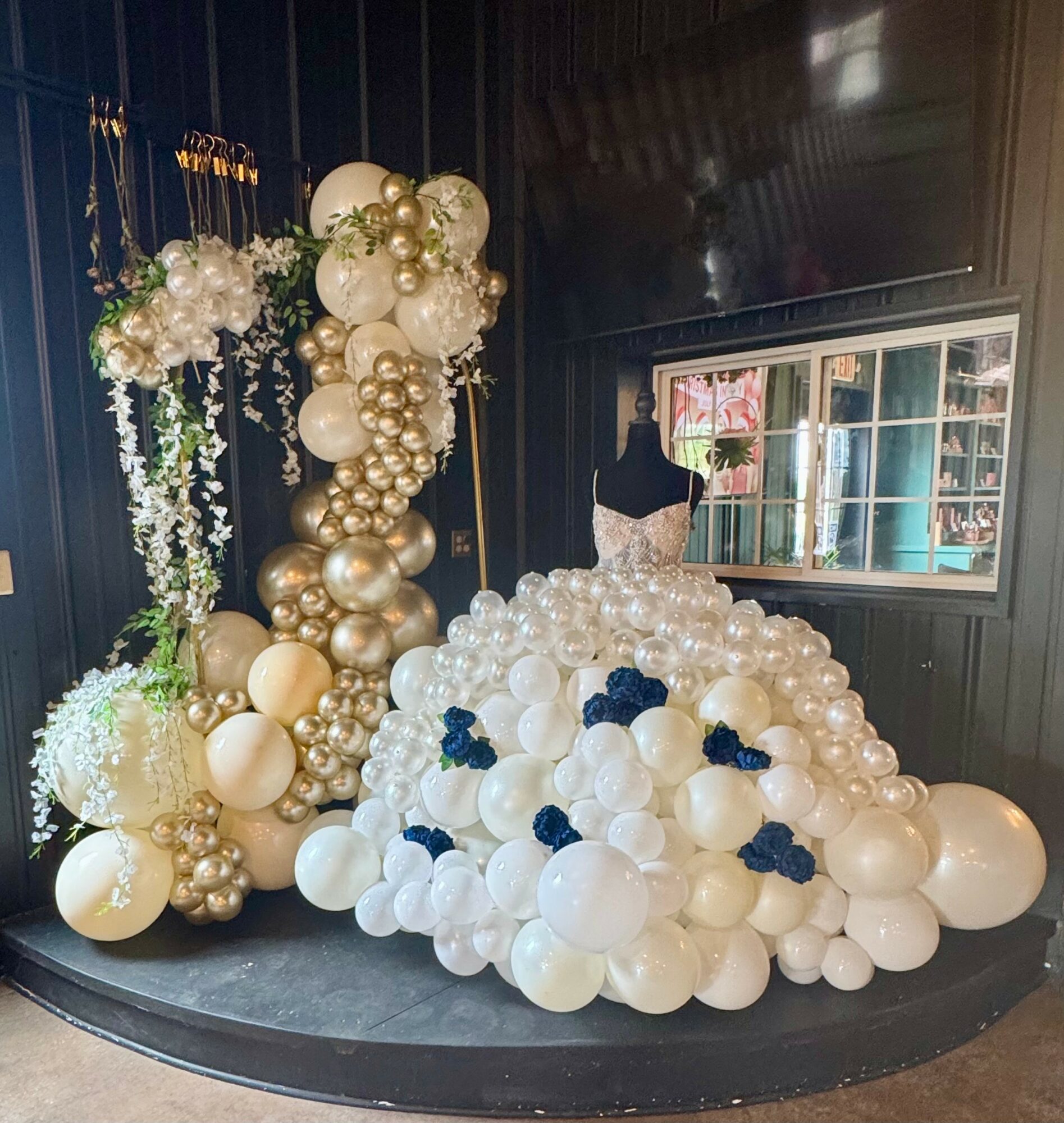 Decorative display with white, gold, and navy balloons arranged in a floral and cascading style, inside a room with a window.