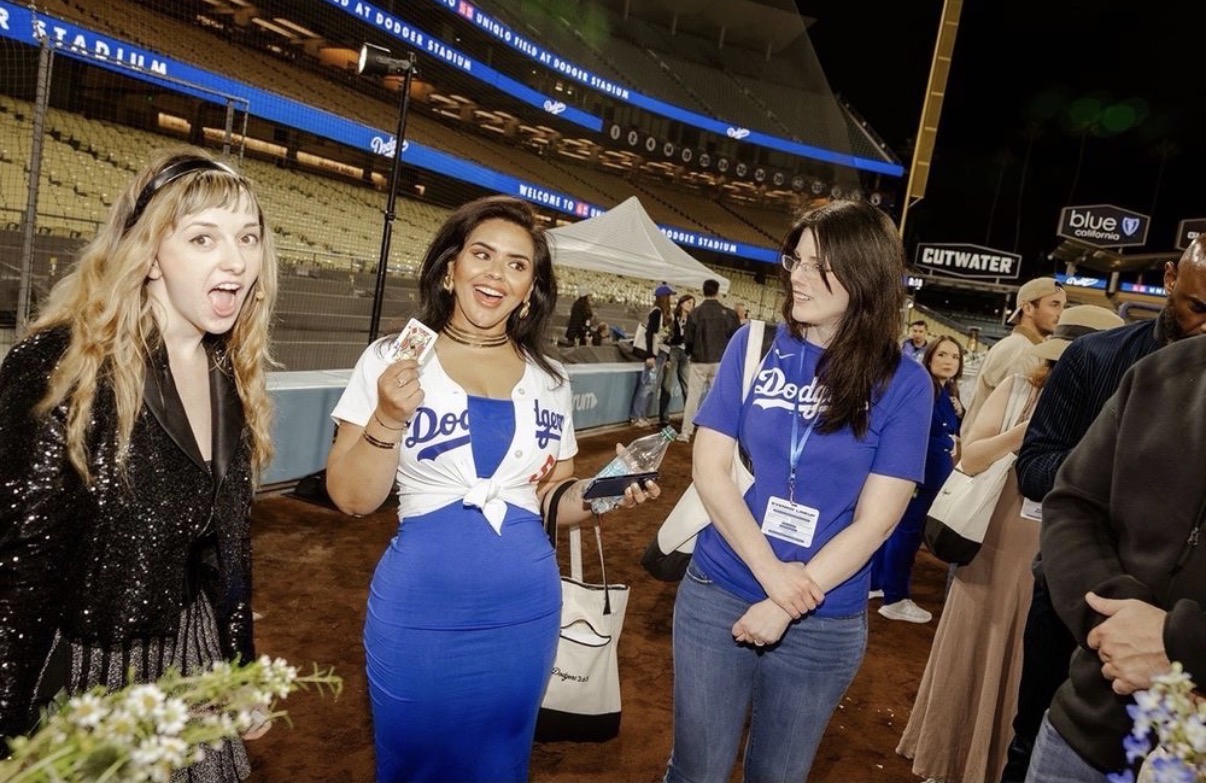 Three women in Dodgers shirts talking at a stadium event, with tents and people in the background.