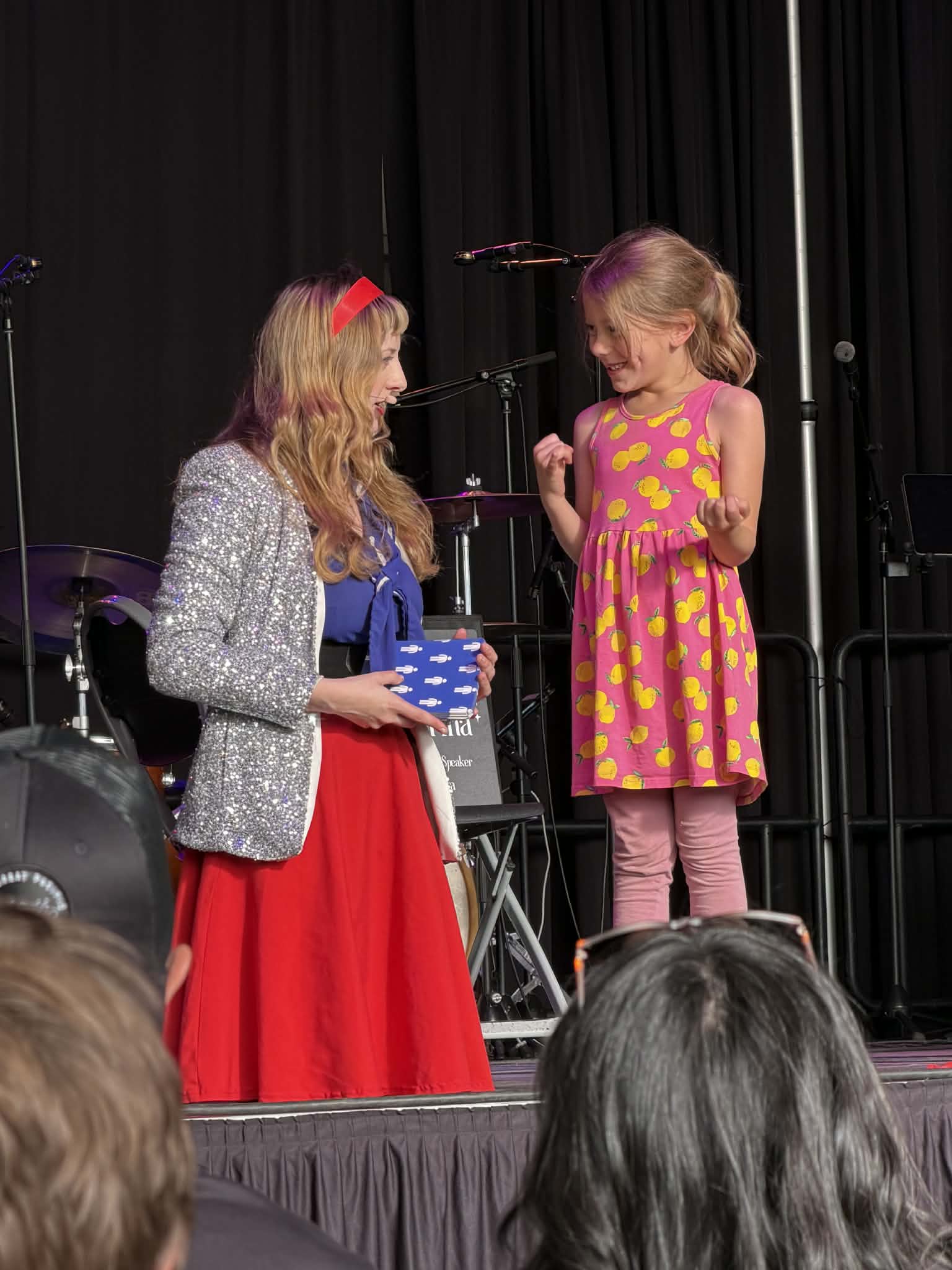 Two girls on stage, one holding a gift box, with a black curtain background and audience in foreground.