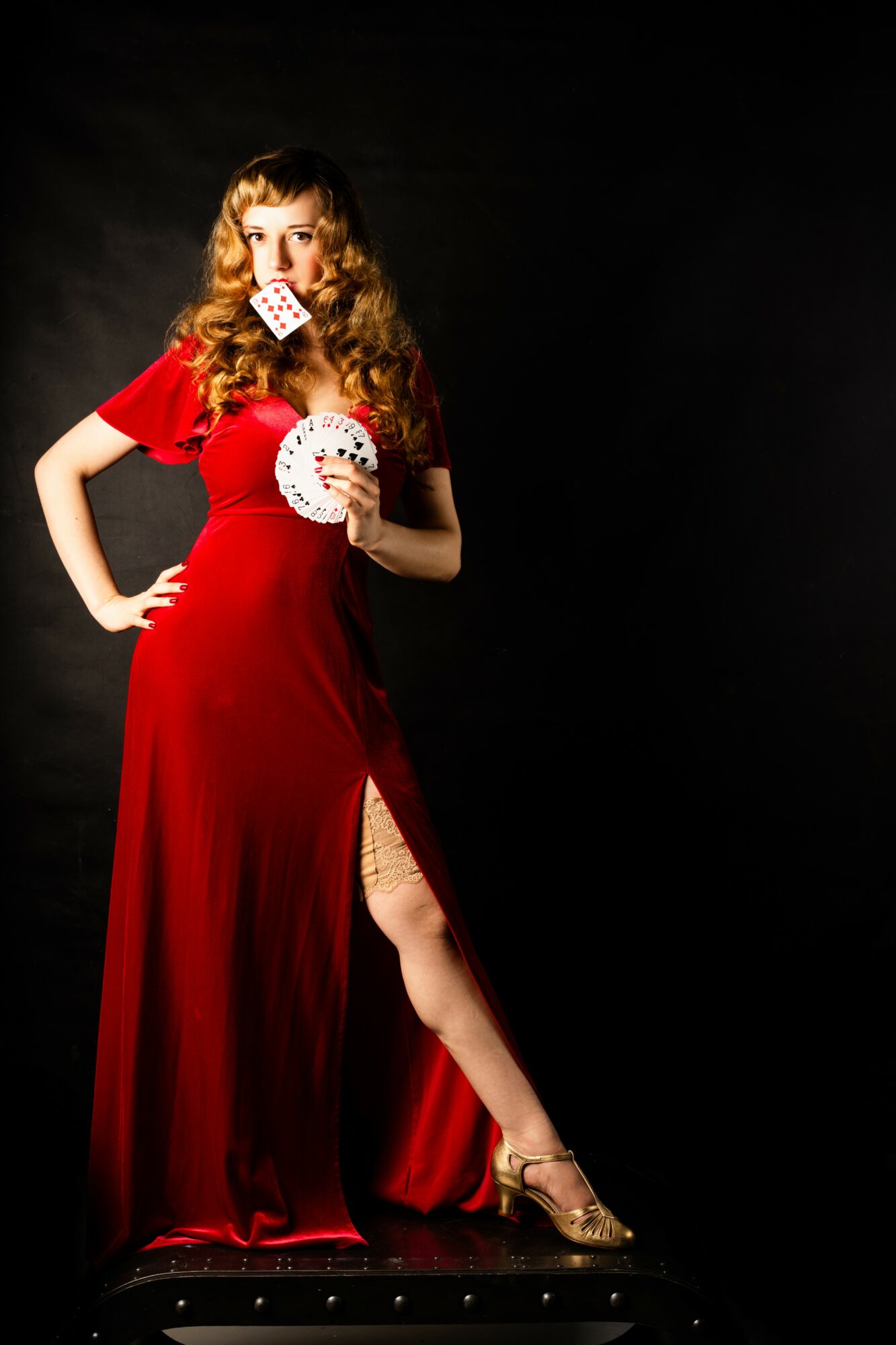 Woman in red dress holding playing cards, standing on a black platform against a dark background.