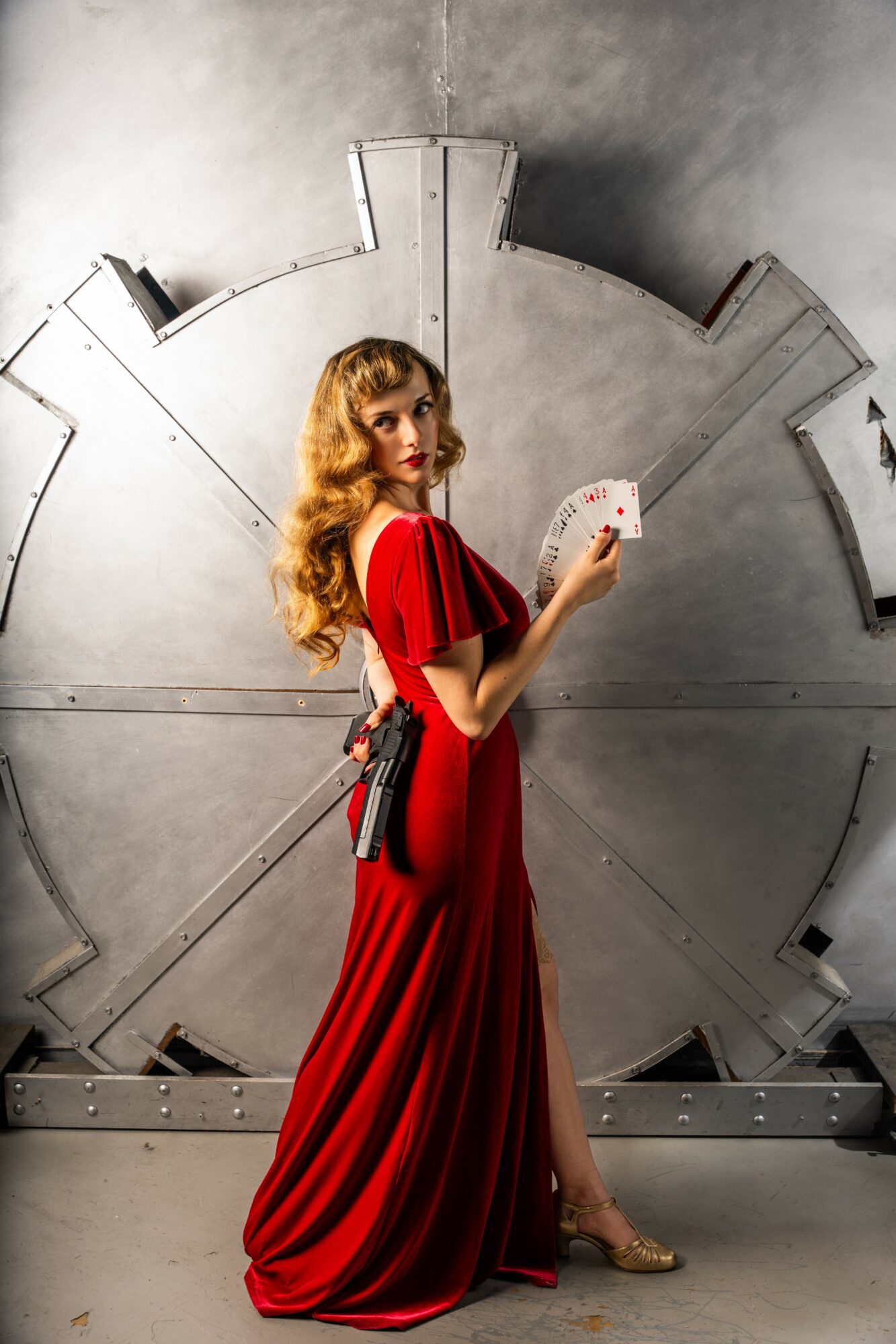 Woman in red dress holding playing cards, standing in front of a large metallic clock face, looking at camera.