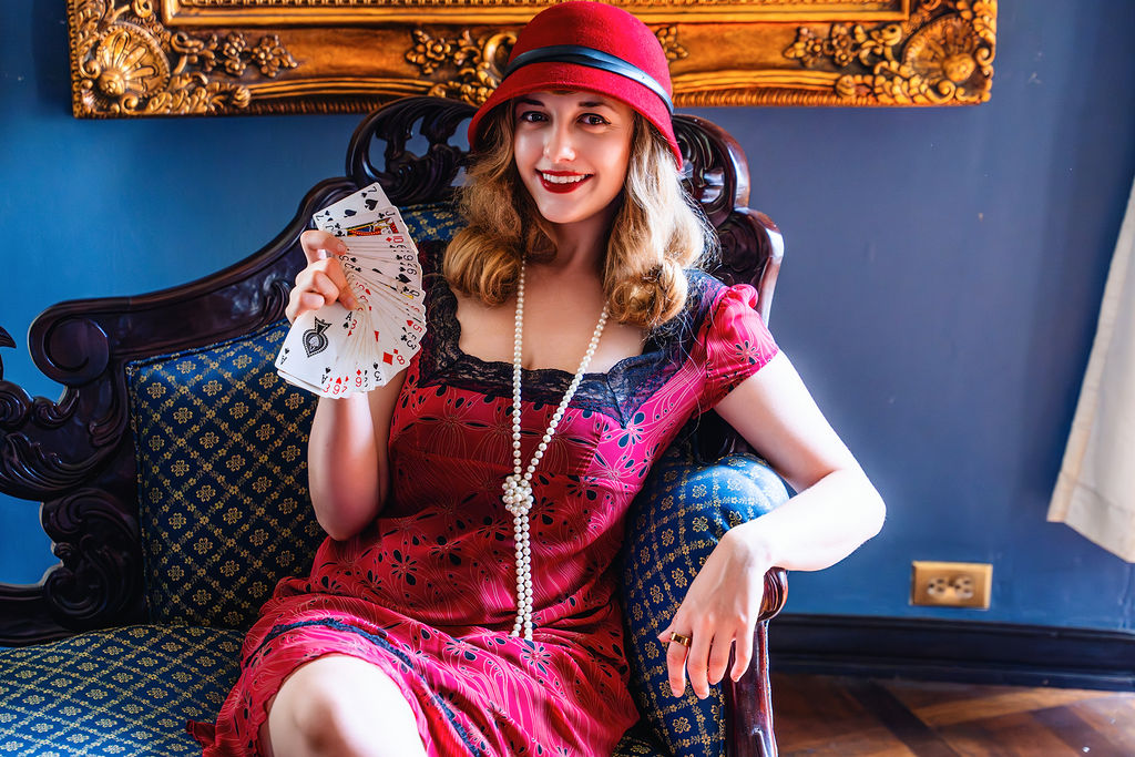 Young woman in vintage dress and red hat sitting on a dark ornate sofa, smiling, holding a phone, in a decorated room.