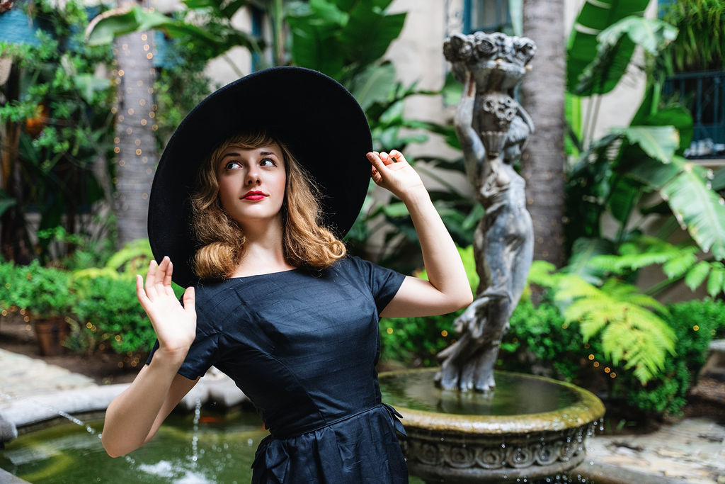 Young woman with wavy hair wearing a large black hat and dark dress, standing near a fountain in a garden.