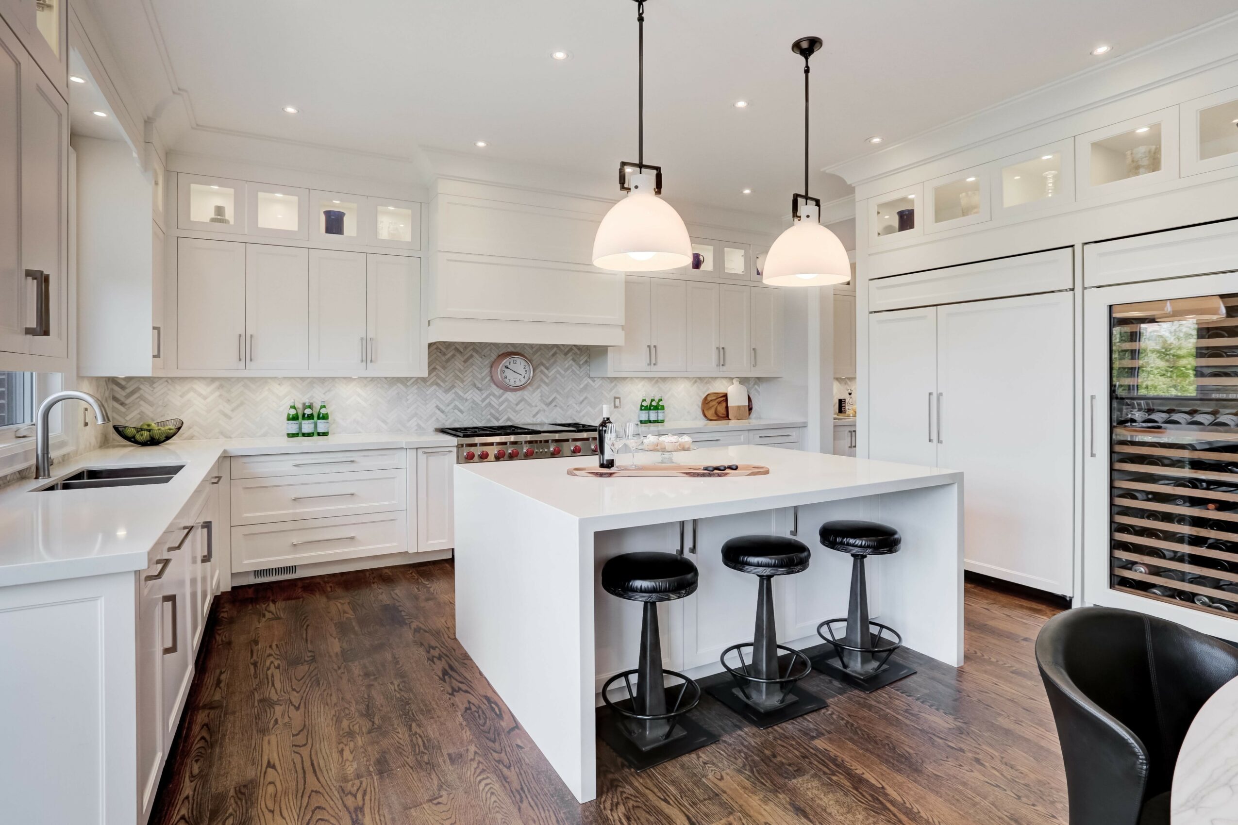 Bright kitchen with white cabinets, island with three black stools, and two pendant lights, hardwood floor, and built-in appliances.