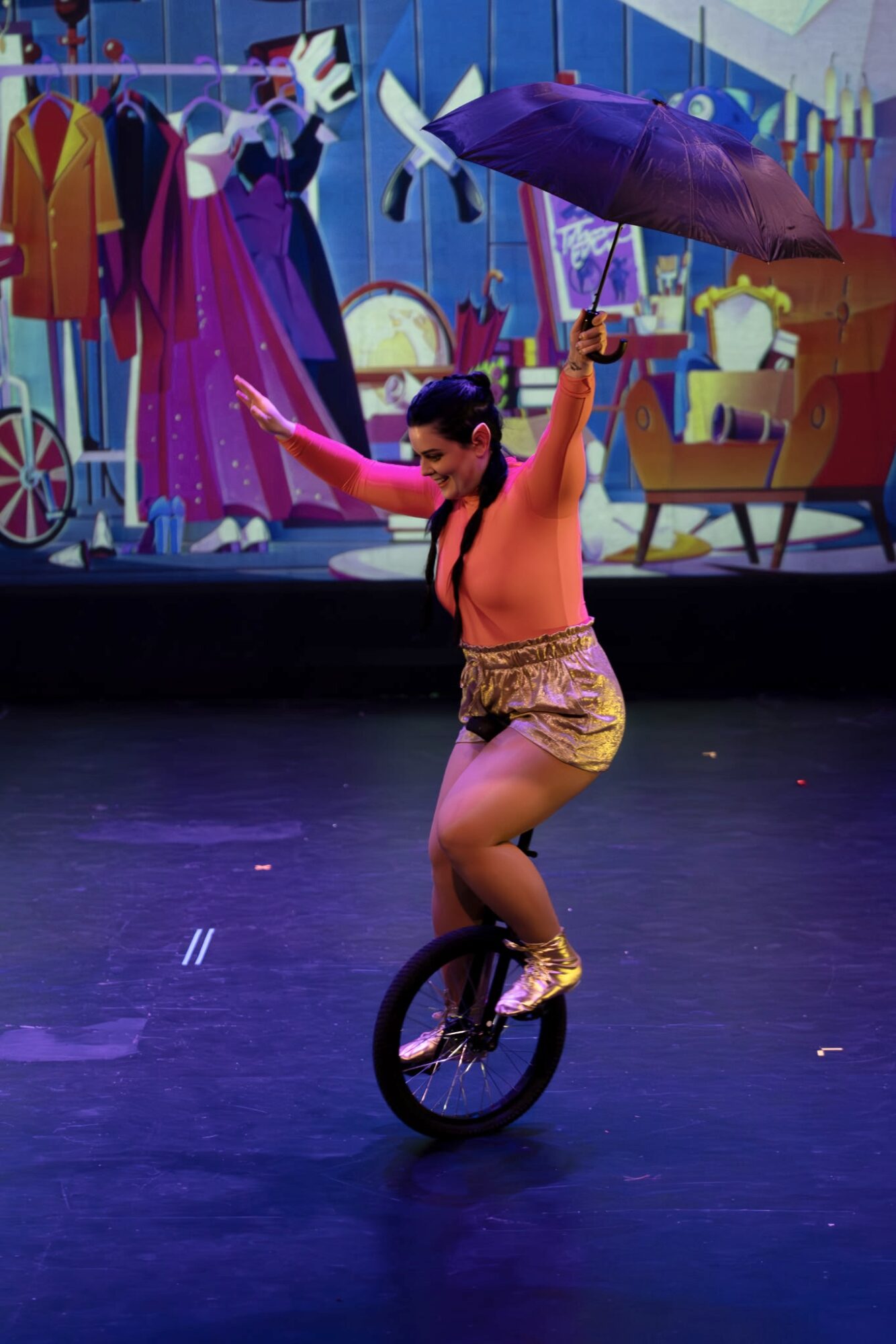 Person balancing on a unicycle, holding an umbrella, with colorful costumes and furniture in background.