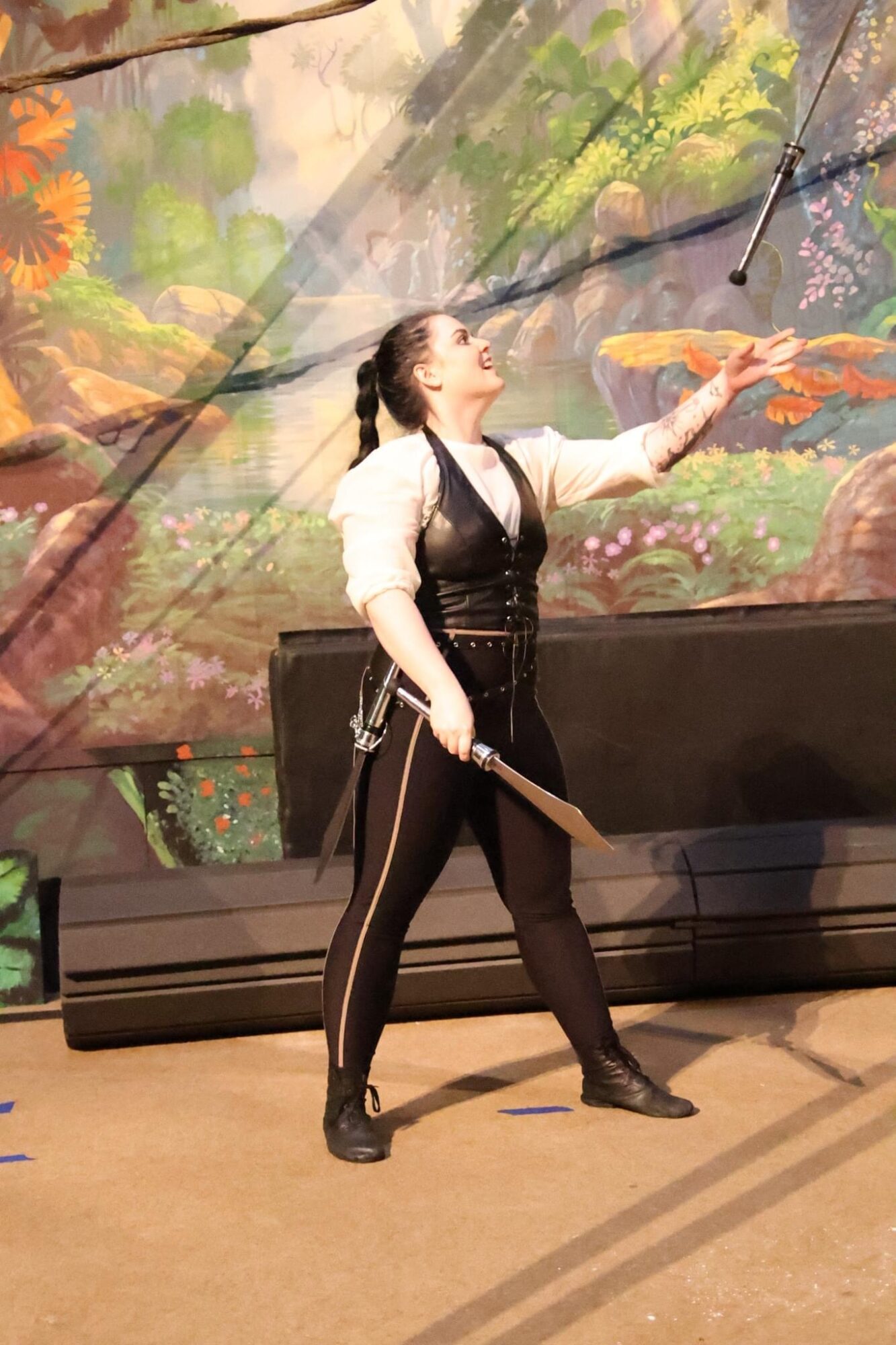 Person performing on stage with a colorful forest backdrop, holding a stick, reaching upward, wearing black pants and a white top.