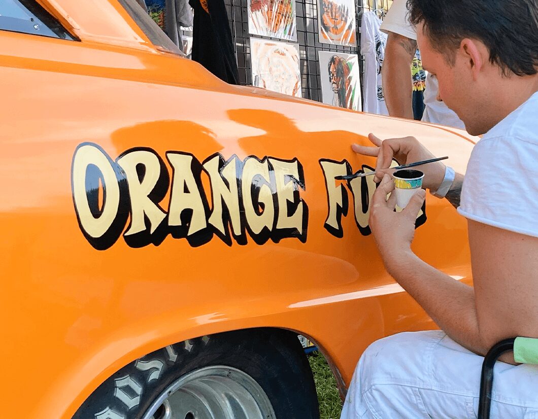 Person painting orange text on a vehicle, with a background of artwork and people, outdoors.