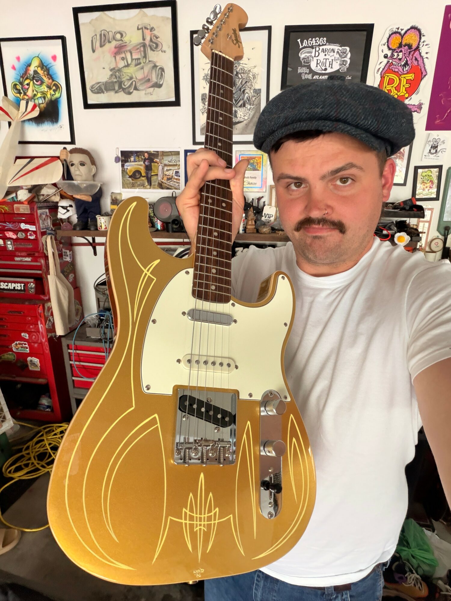 Person holding a gold-colored electric guitar in a room with framed pictures and shelves.