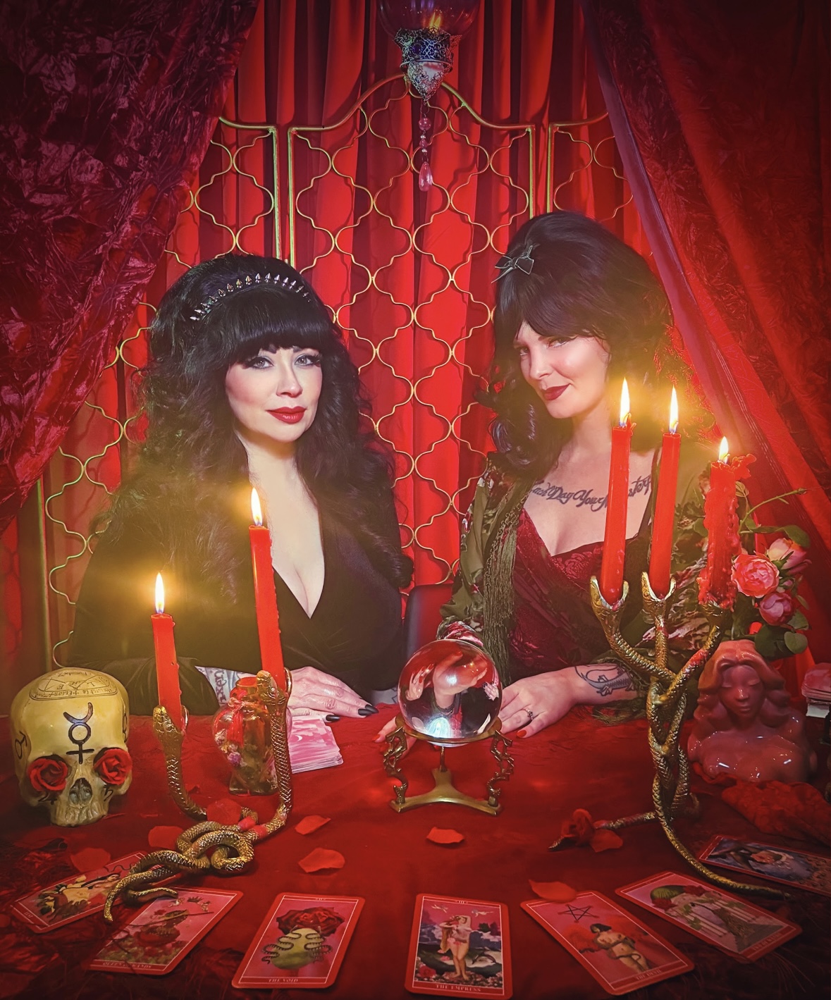 Two women with dark hair and makeup sit at a table with candles, tarot cards, and decorative items, against red curtains.
