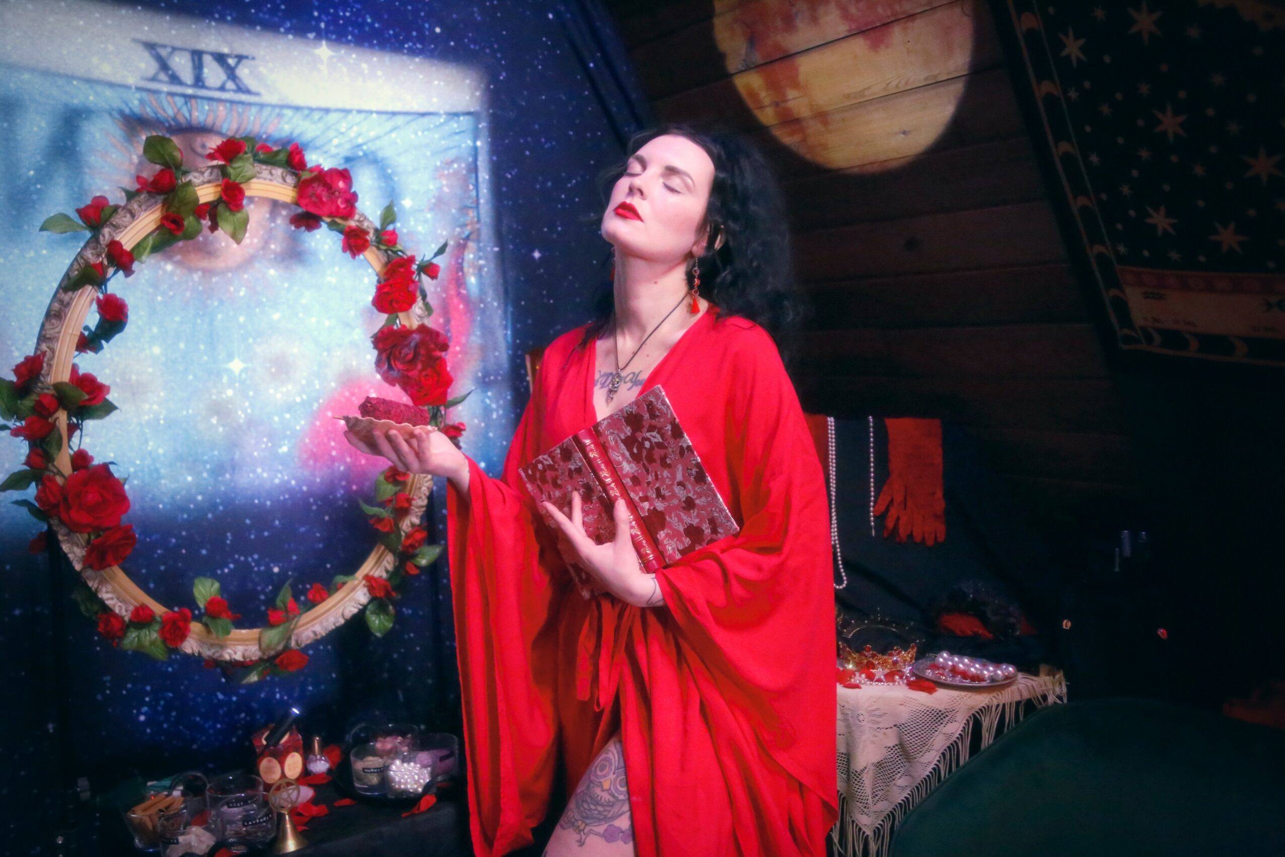 Woman in red dress with closed eyes, holding a book, standing near a decorated window with a wreath, in a cozy room.