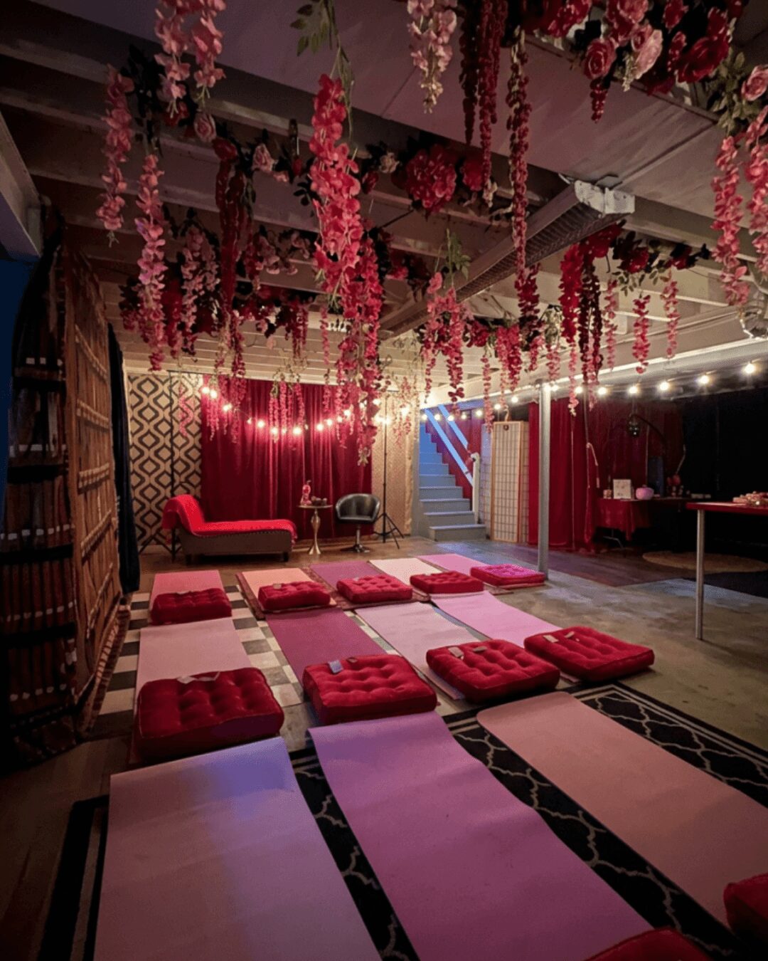 Indoor space decorated with hanging pink flowers, red seating, cushions on the floor, and a stage area with curtains.