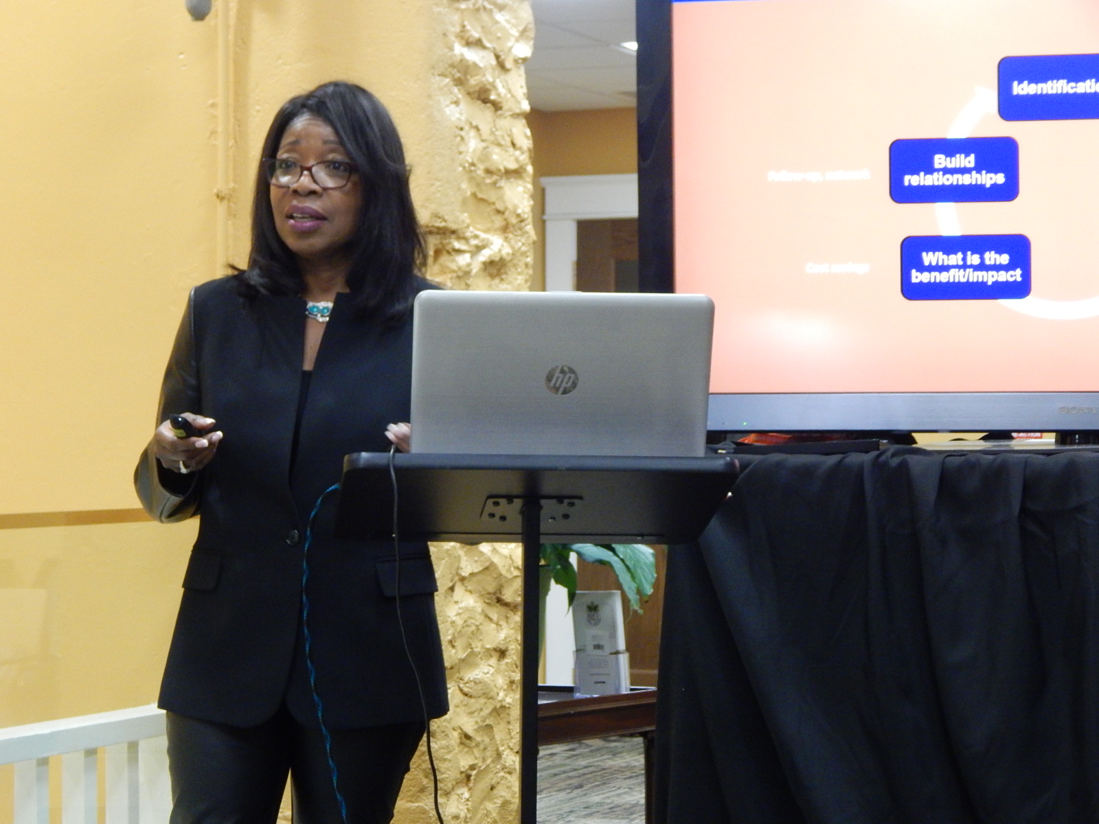 Woman in black blazer presenting with a laptop and a screen displaying a presentation, in a room with yellow walls.