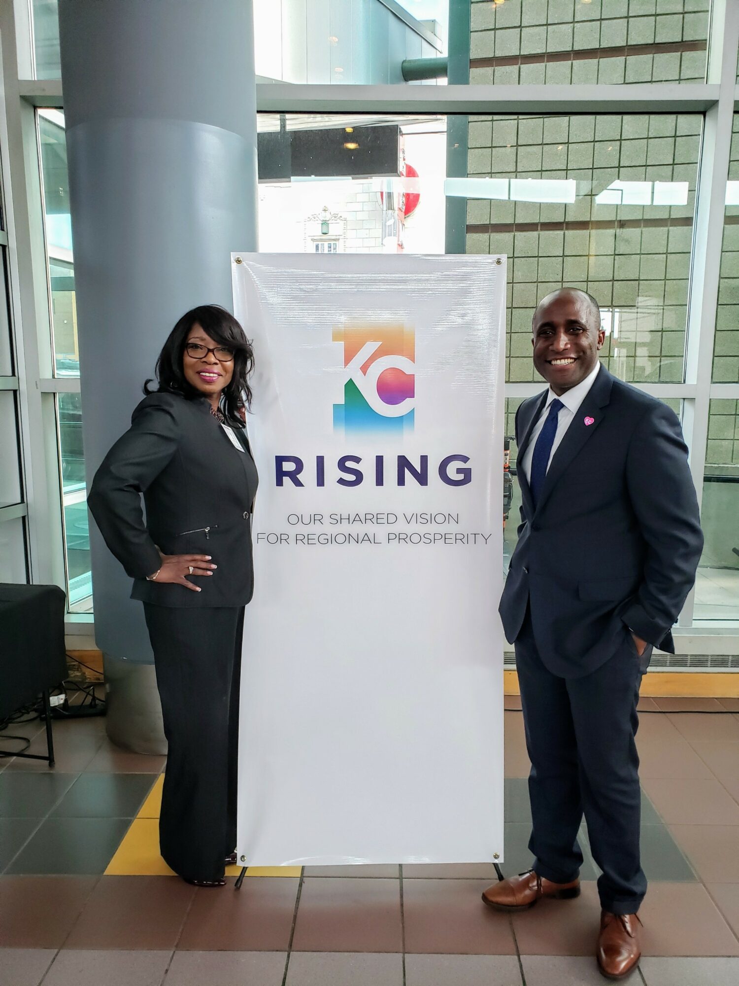 Two people stand on either side of a vertical banner in a modern building with large windows, smiling at the camera.